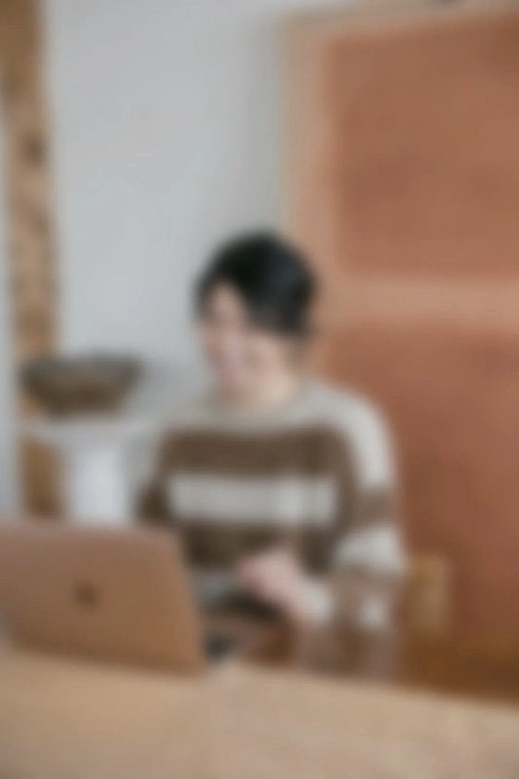 A person with short dark hair wearing a striped shirt, sitting at a desk with a laptop in front, in an indoor setting with a blurred background.