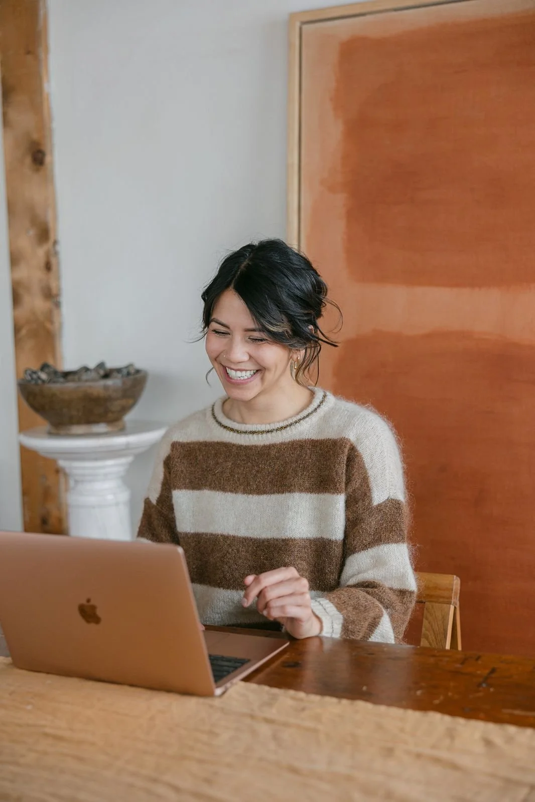 A woman with short dark hair wearing a beige and brown striped sweater, smiling and looking at her open pink MacBook on a wooden table.