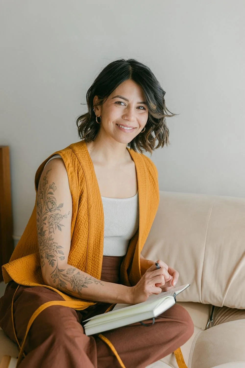 A woman with dark wavy hair, earrings, and a nose ring sitting on a beige couch, smiling, wearing a white top, orange vest, and brown pants, holding a pen and a notebook.