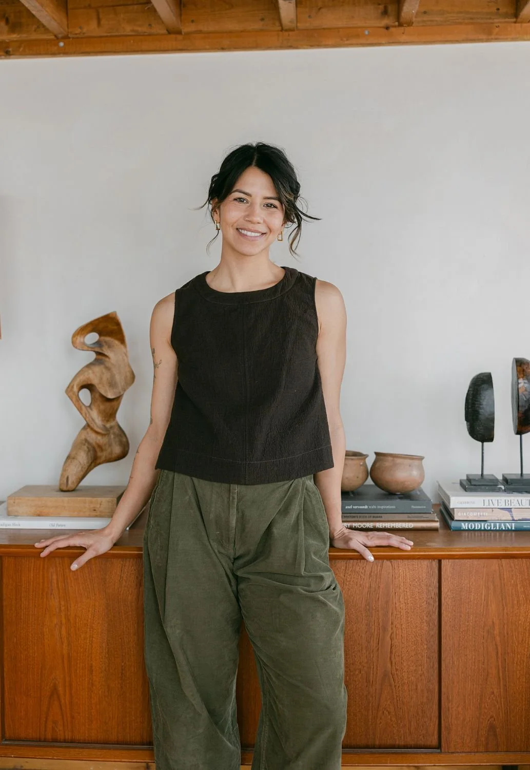 A woman with shoulder-length black hair, wearing a sleeveless black top and olive green pants, standing in front of a wooden sideboard decorated with art pieces, books, and pottery in a room with a white wall and wooden ceiling.