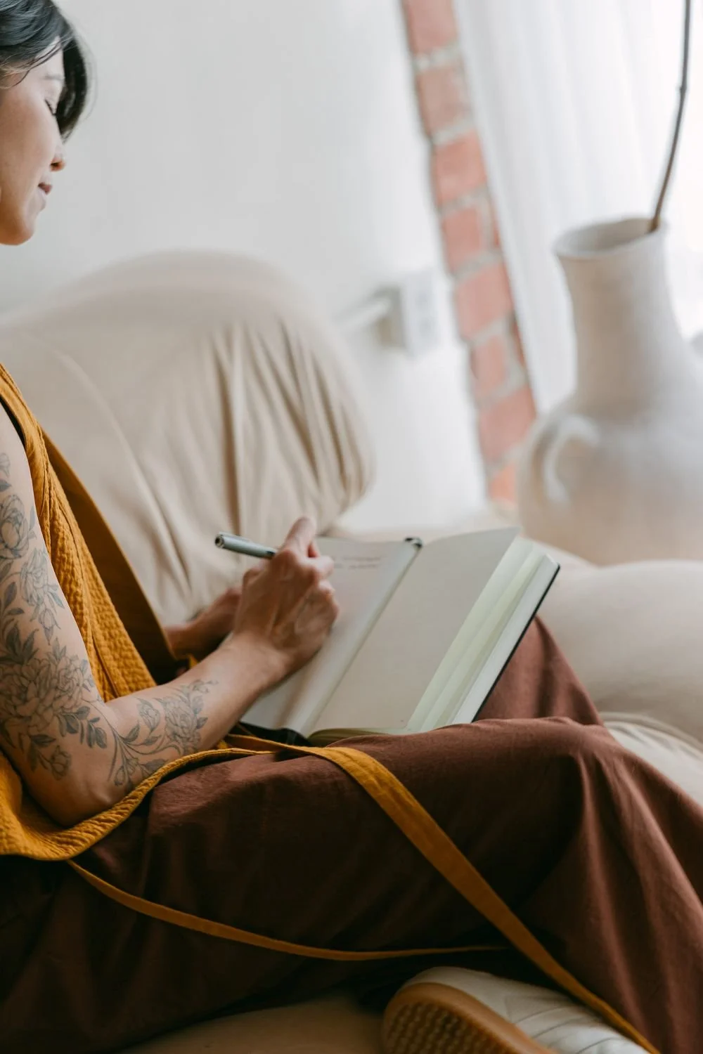 A woman with tattoos sitting on a beige couch, writing in a notebook with a pen, wearing a mustard yellow sleeveless top and brown pants, next to a white brick wall and a large white ceramic vase.