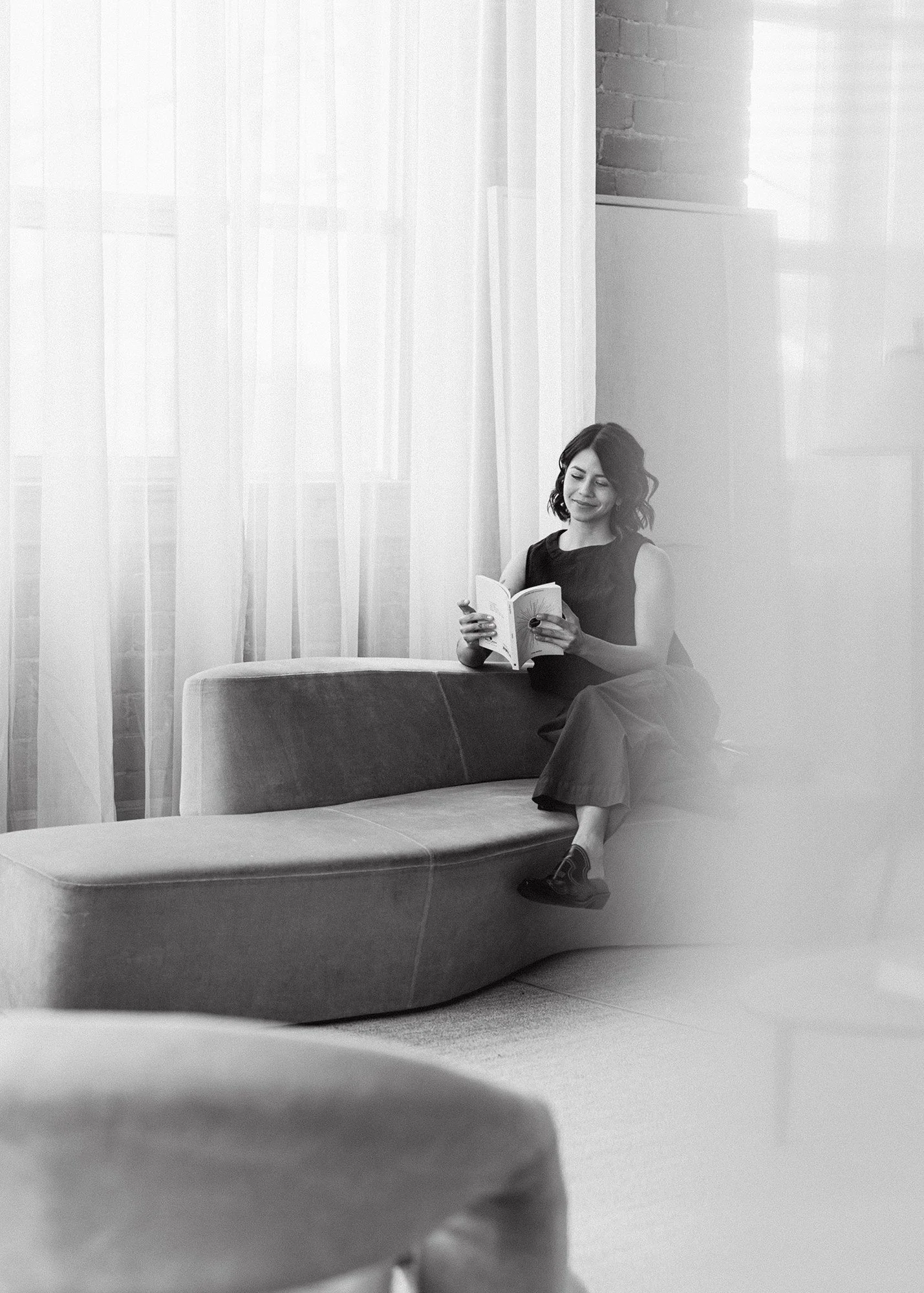 A woman with shoulder-length hair, wearing a sleeveless top and wide-legged pants, sits on a modern curved sofa, reading a book in a bright room with sheer curtains and brick wall accents.