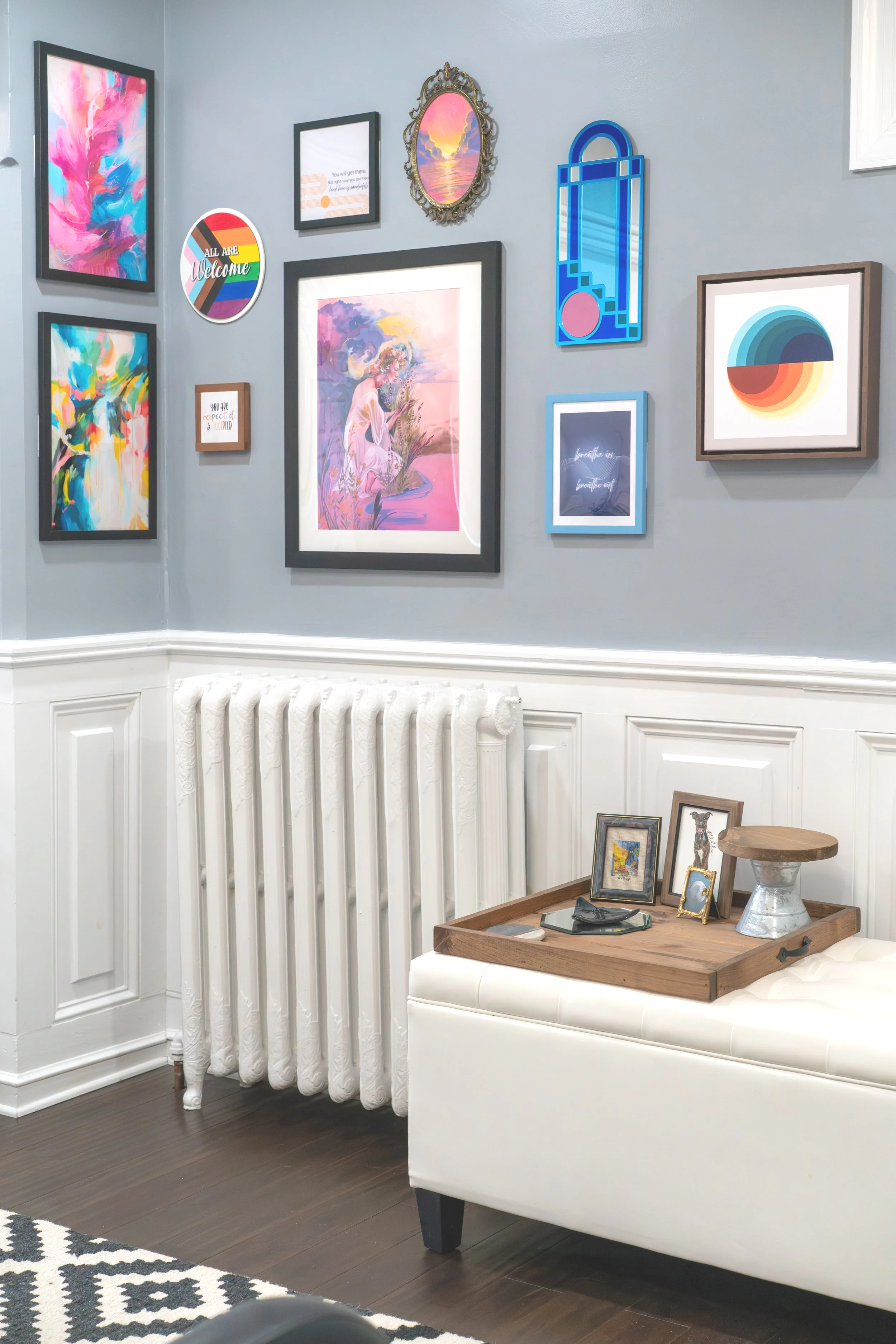 A corner of a room with a gallery wall of colorful and abstract artwork, a white radiator, a small white tufted bench with a wooden tray on top holding framed photos and decor, and a section of a patterned black and white rug.