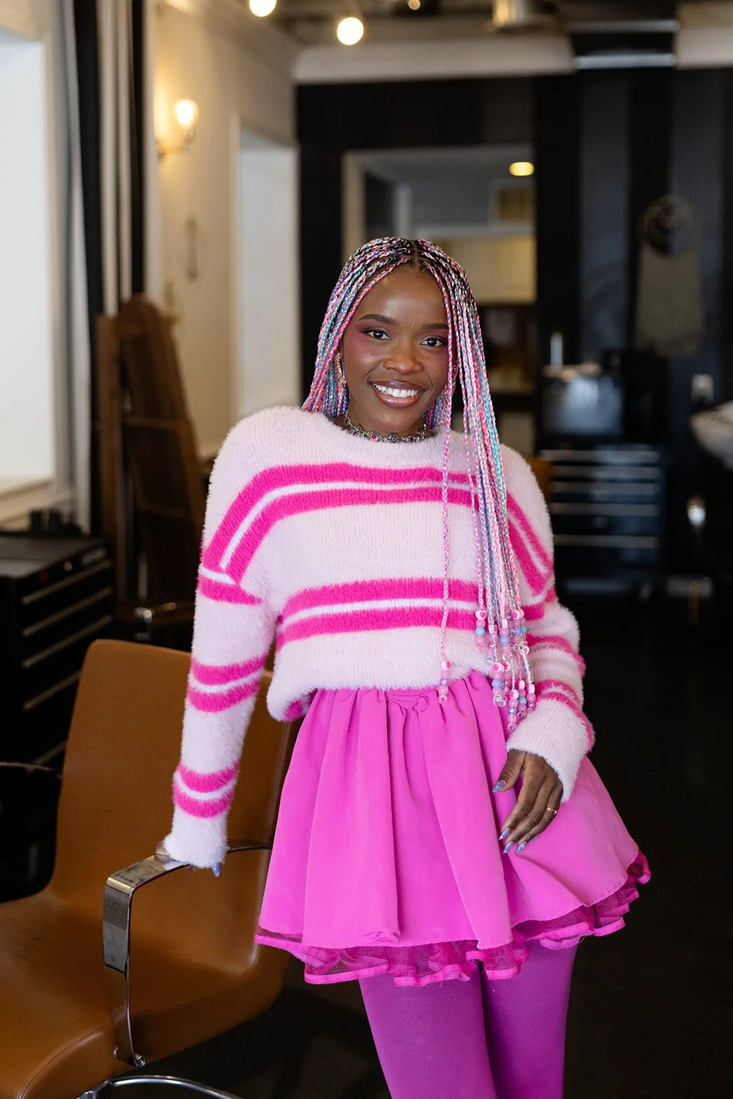 A woman with short, colorful hair dyed in teal, purple, and pink, smiling and wearing bright makeup and jewelry. She has vibrant blue lipstick, pink and purple eye makeup with decorative rhinestones, and a printed pink dress with artistic illustrations.