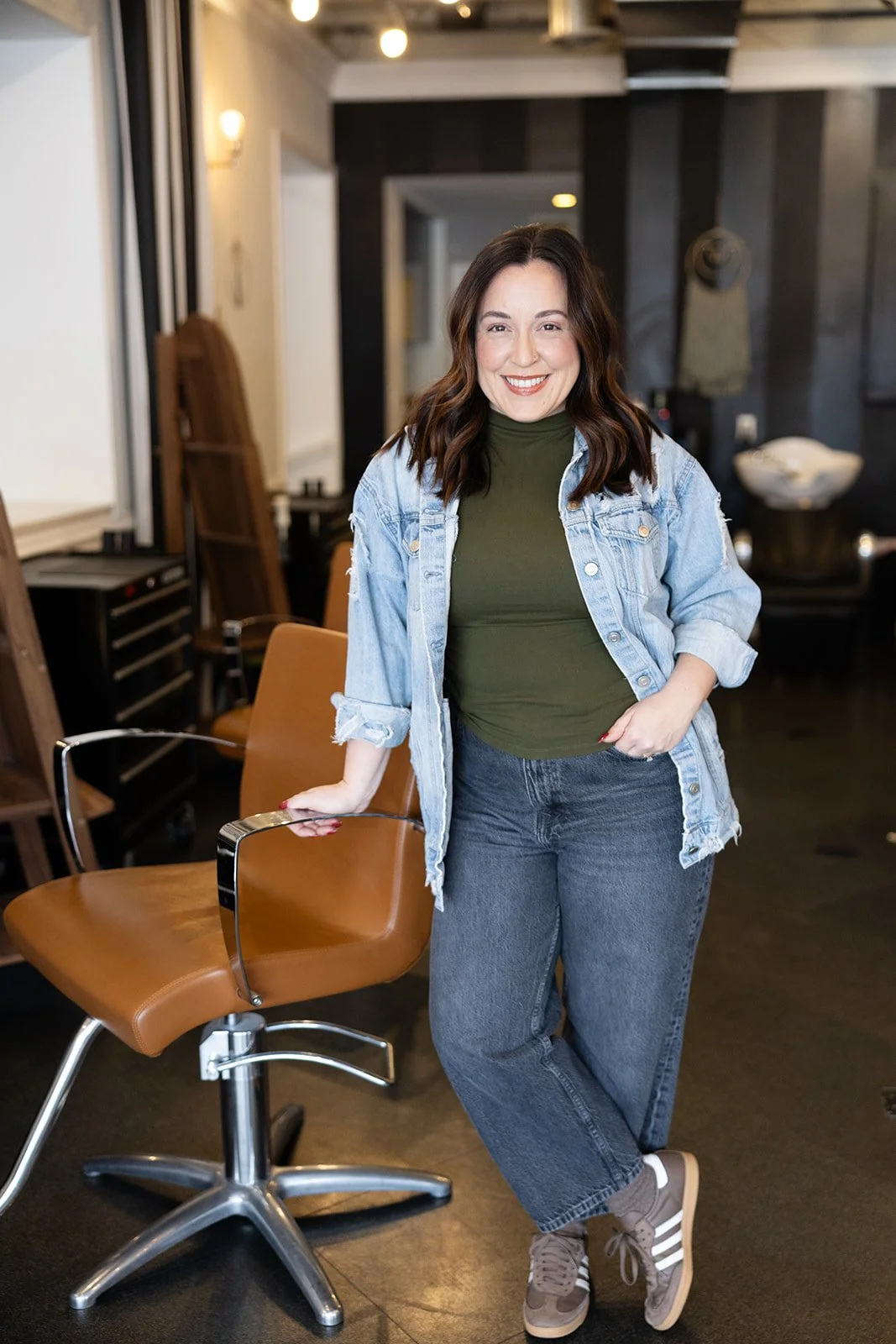 Smiling person with dark hair, wearing a camouflage jacket and necklace, in front of a plain background.