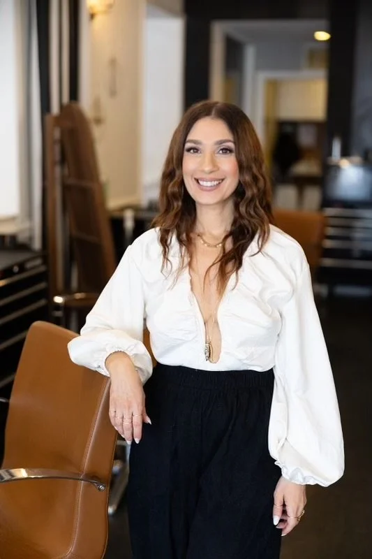 A woman with wavy brown hair and a wide smile, wearing a white blouse and a gold necklace, standing against a plain light-colored background.