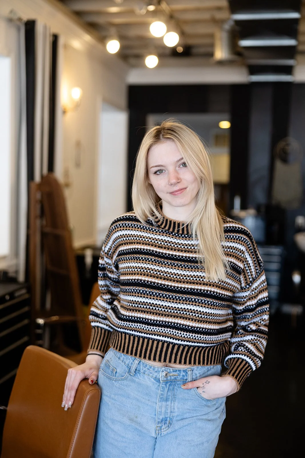 A young woman with shoulder-length brown hair, hazel eyes, and a septum piercing, smiling softly while wearing a green lace-trimmed tank top, standing against a light-colored wall.