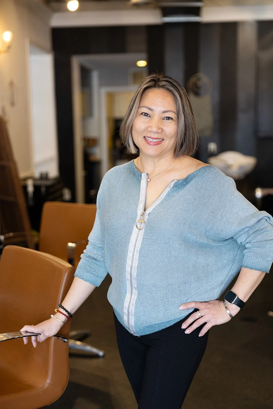 A smiling woman with short, light brown and blonde hair wearing a black top and a thin necklace, standing against a plain light-colored background.