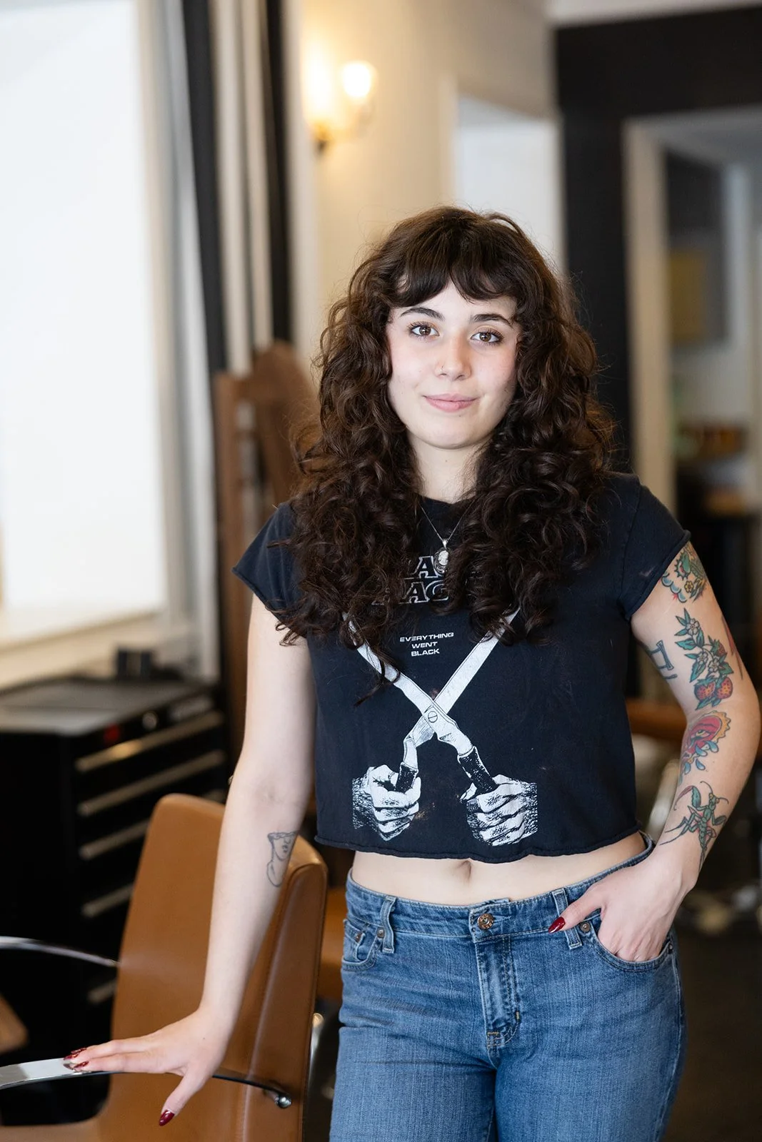 Person with curly hair, red lipstick, wearing a black dress and jewelry, smiling at the camera.