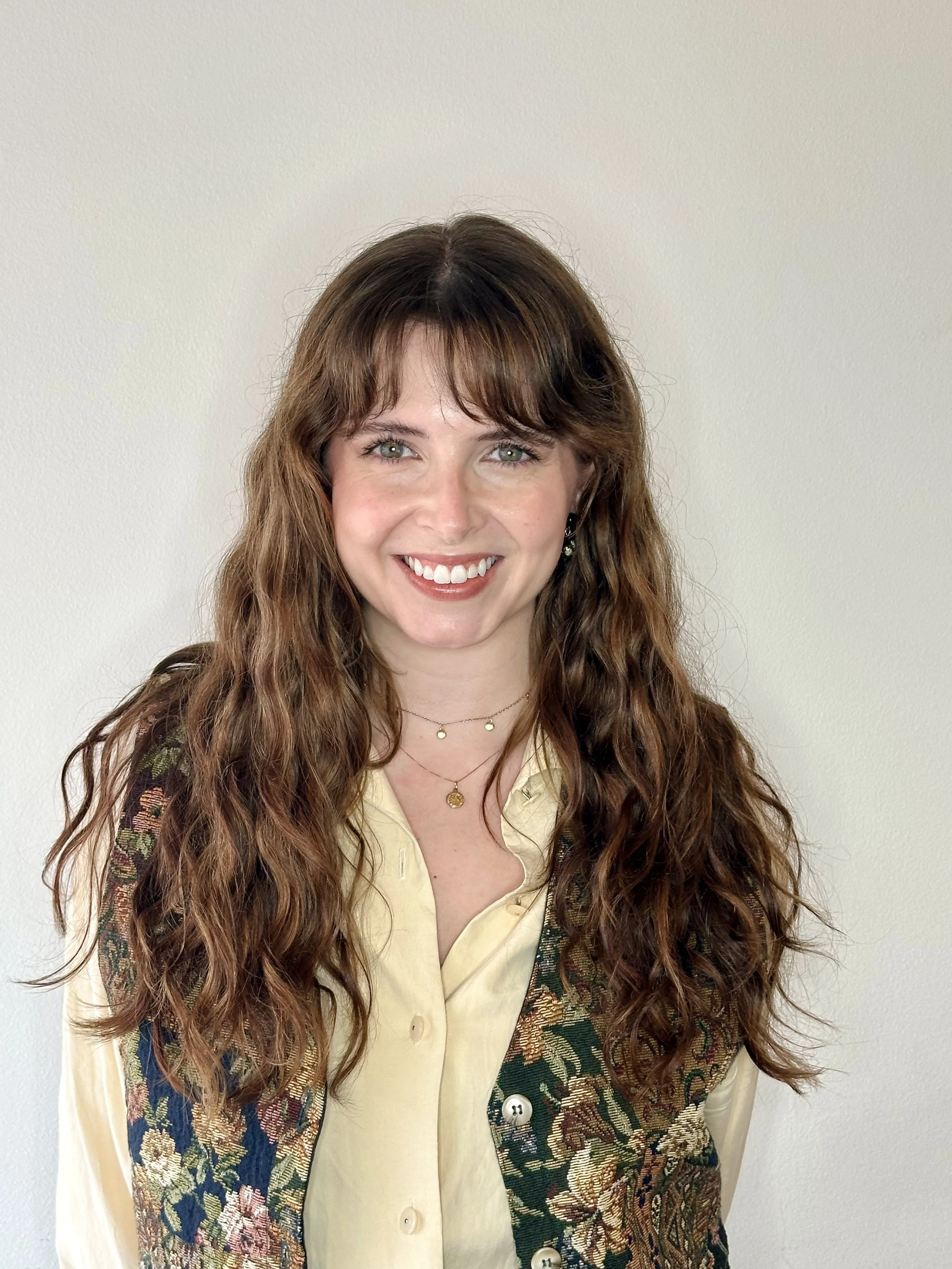 A woman with red hair and a cheerful smile, wearing a cream turtleneck sweater and a beige and yellow headscarf, touching her hair.