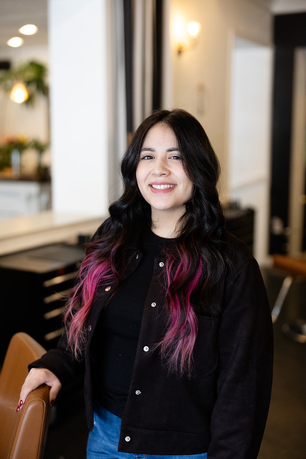 A young woman with dark, wavy hair smiling and holding her hands in her hair, wearing a black t-shirt, standing against a plain light-colored wall.