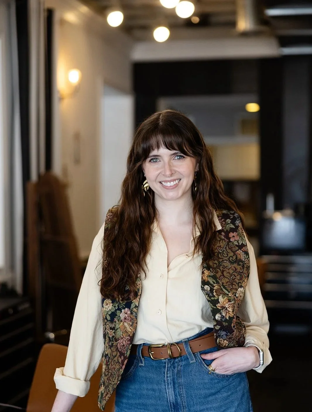 A woman with red hair and a cheerful smile, wearing a cream turtleneck sweater and a beige and yellow headscarf, touching her hair.