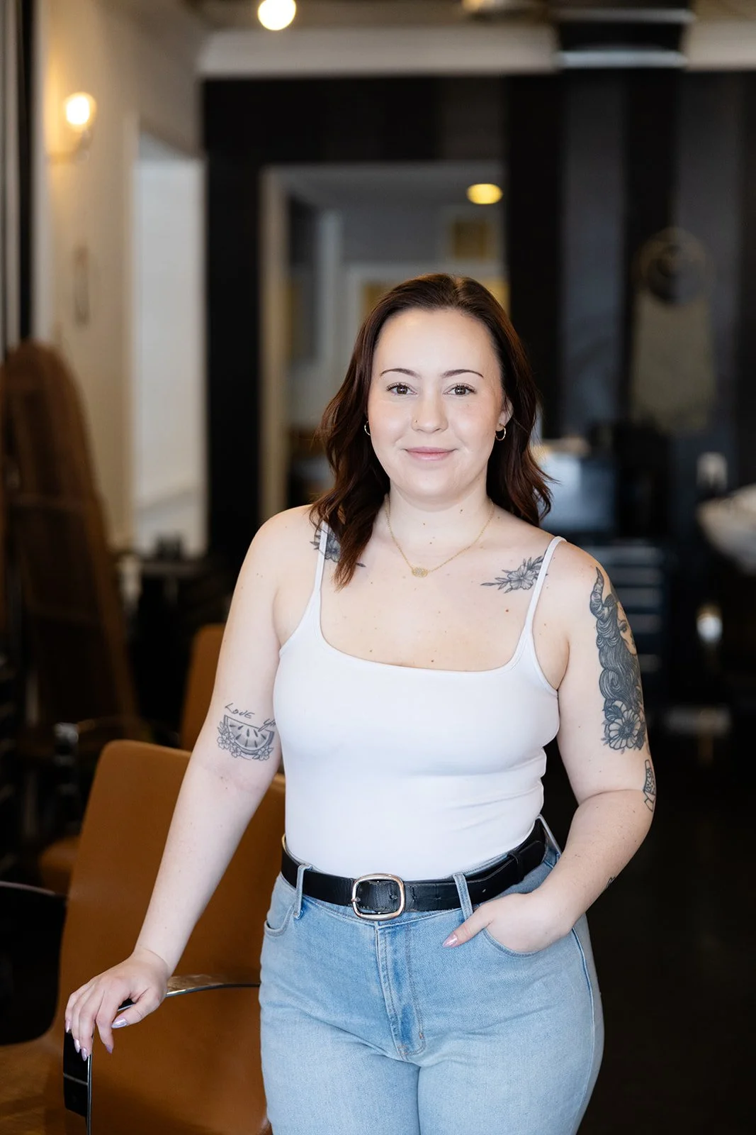 Portrait of a smiling woman with shoulder-length brown hair, wearing a red floral dress with thin straps, and layered necklaces, standing against a plain light-colored wall.