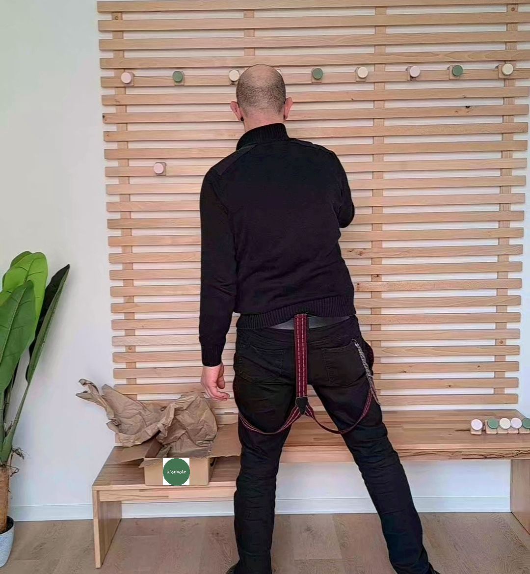 Ein Mann steht mit dem Rücken zur Kamera vor einer vorgezeichneten Holzwand und trägt eine Sicherheitsleine. Es befinden sich Grüne, Rosa und Weiße Kreise auf der Wand und auf dem Boden. Es gibt eine grüne Pflanze auf der linken Seite.