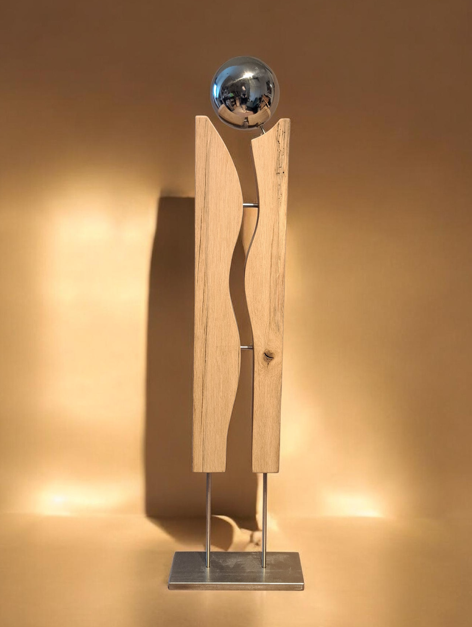 Moderner Skulptur aus Holz, Metall und einer reflektierenden Kugel, auf einem rechteckigen Metallboden, vor einem warm beleuchteten Hintergrund.