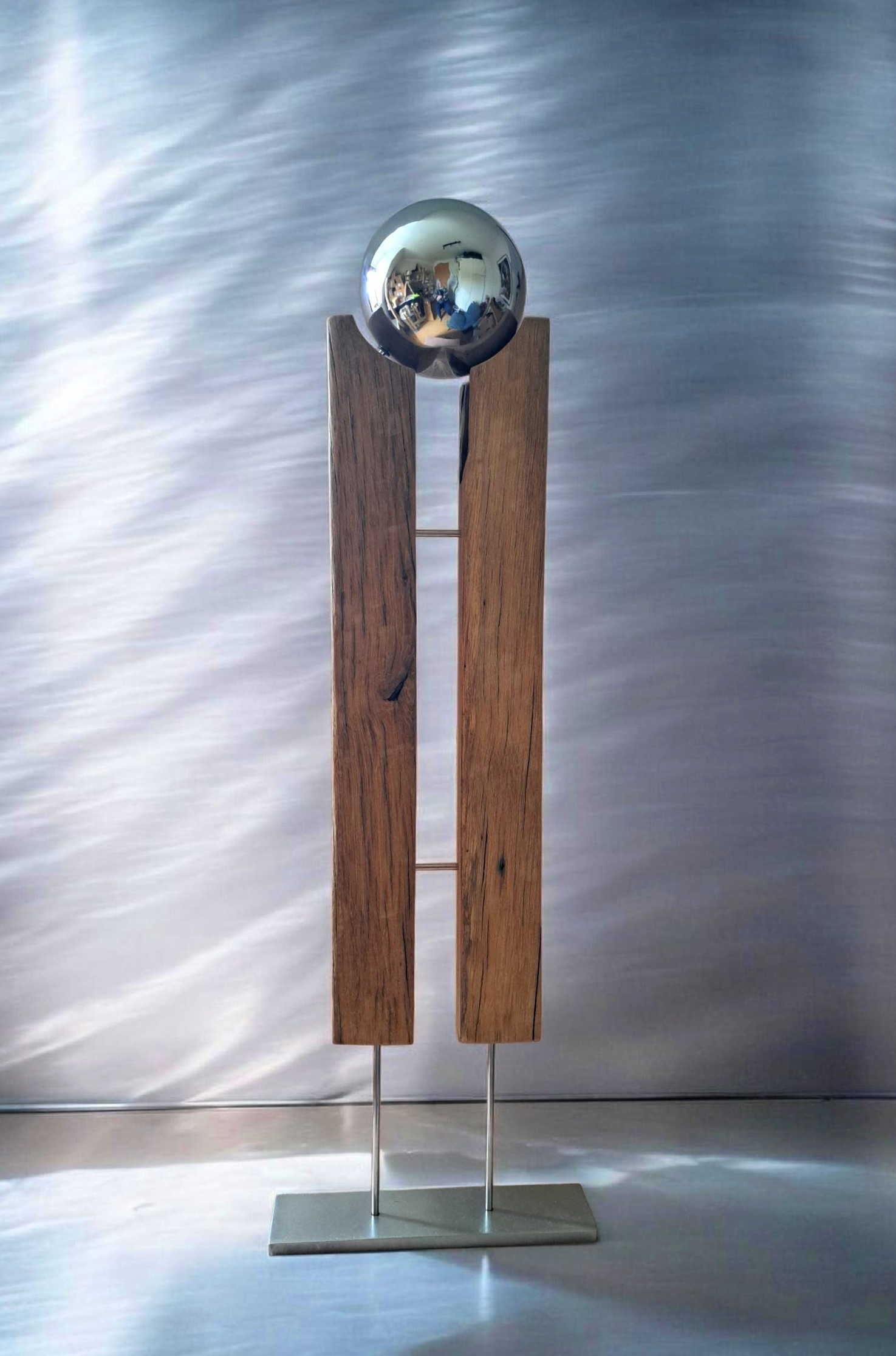 Skulptur besteht aus einem spiegelnden Metallkugel auf zwei vertikalen Holzbalken, die auf einer metallenen Basis befestigt sind.