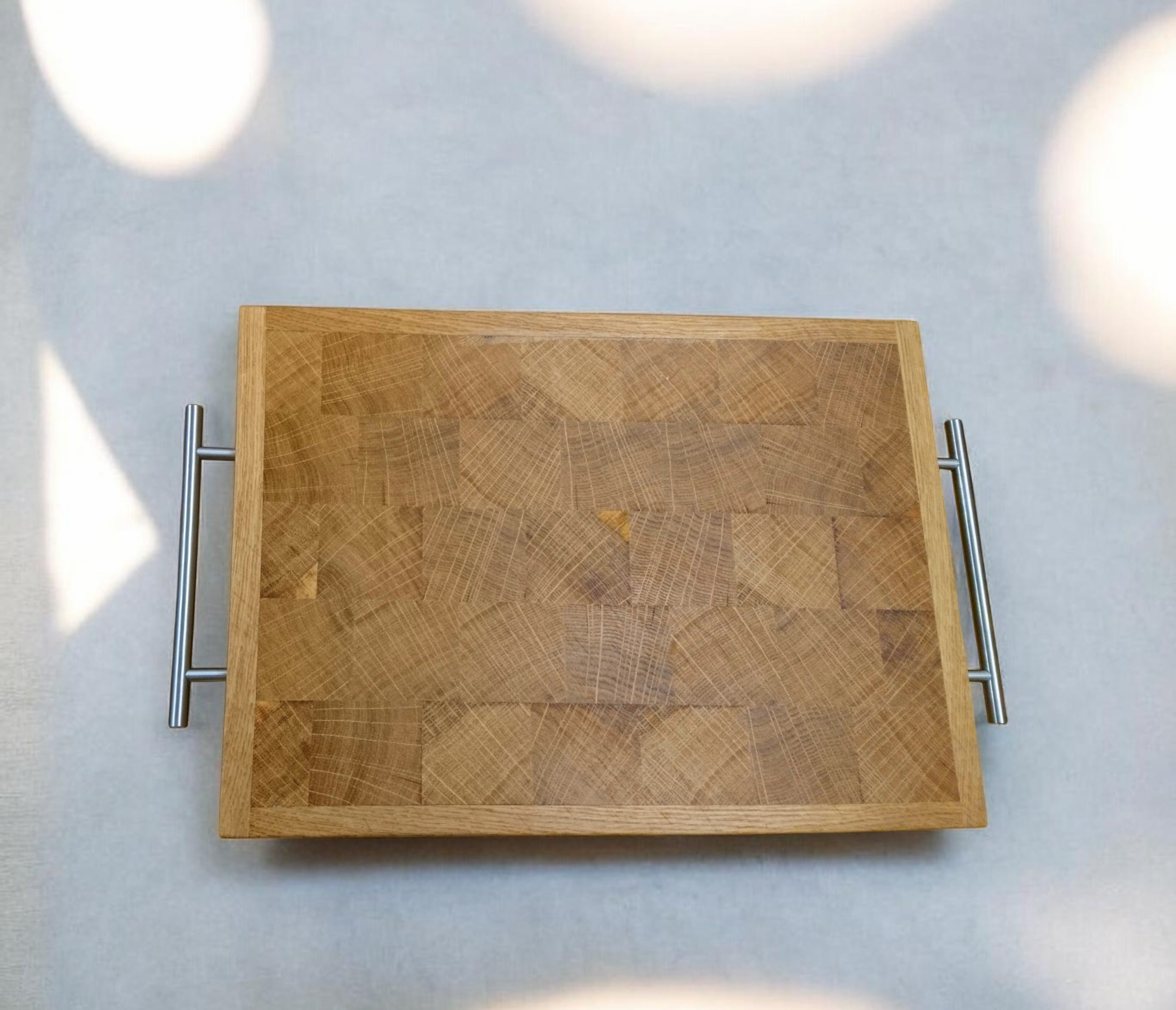 Ein quadratischer Holzbretter mit einem Metallgriff auf beiden Seiten, liegt auf einer hellen Oberfläche und wird von Sonnenlicht und Schatten umgeben.