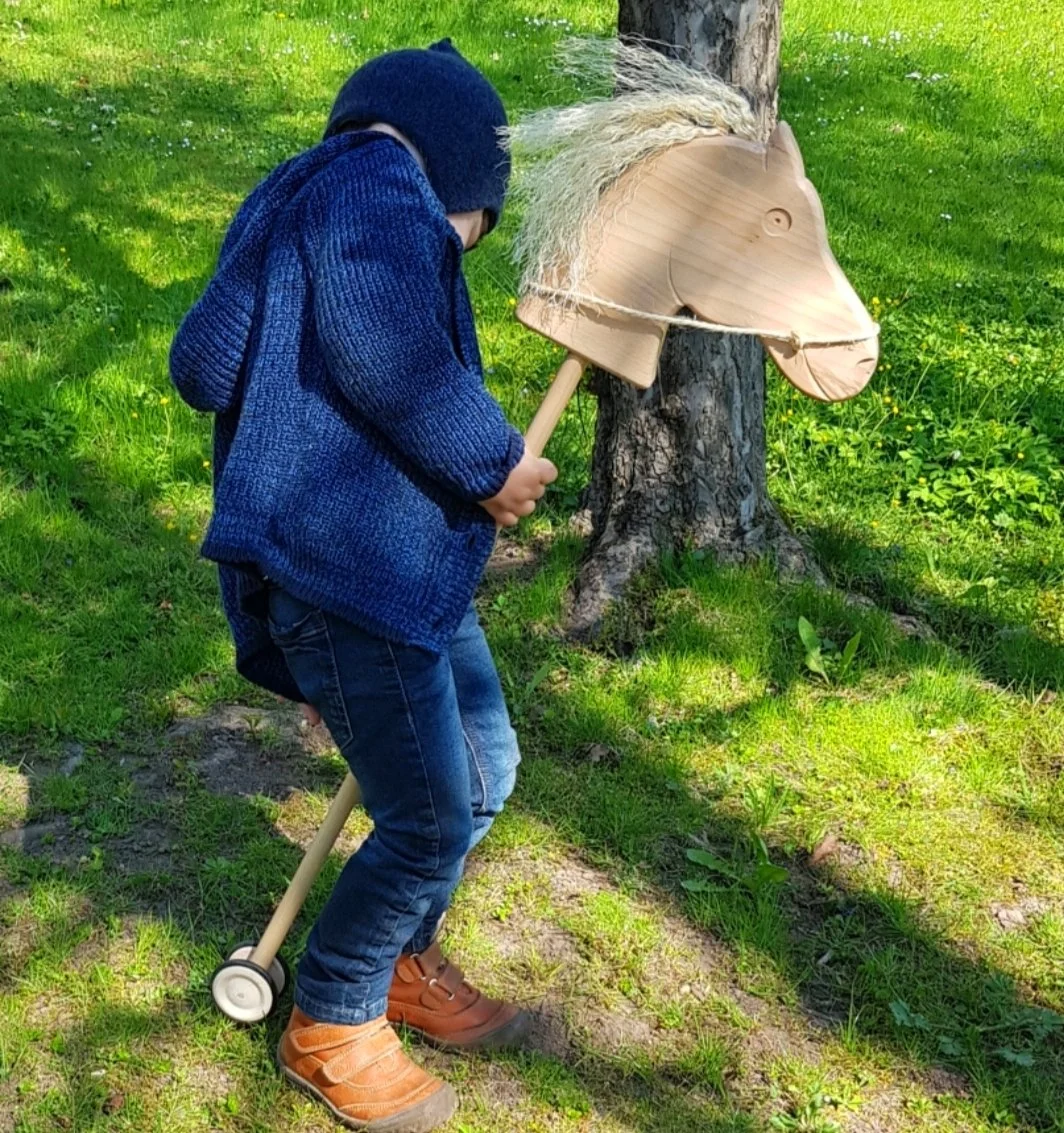 Ein Junge mit blauer Jacke und Jeans spielt im Freien mit einem Holzpferdekopf, der an einem Baum befestigt ist, während er einen Holzstab hält, um das Pferd zu steuern.