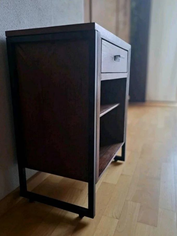 Ein dunkles Holzschränkchen mit einer kleinen Schublade und einem offenen Fach, steht auf einem Holzboden neben einer Wand.