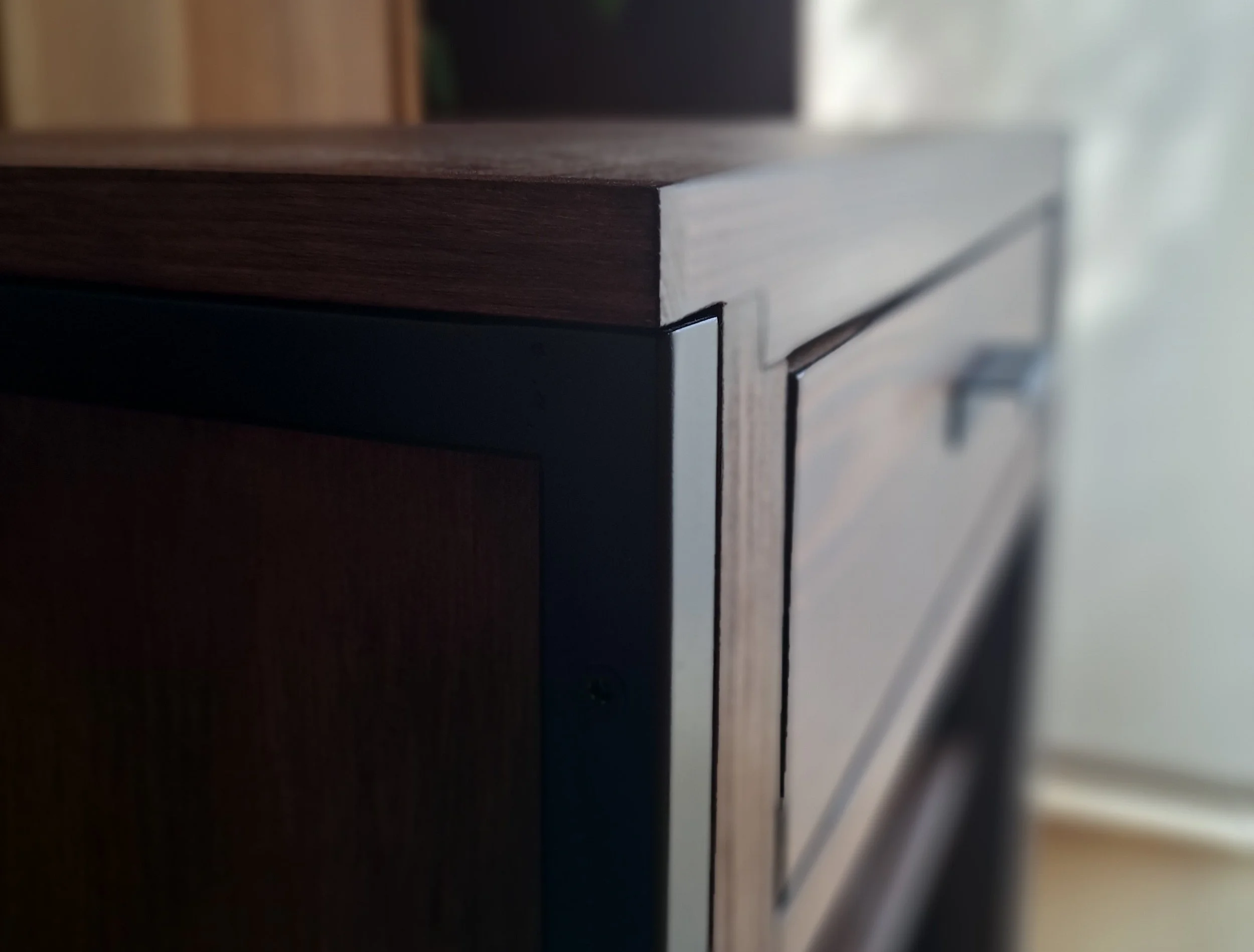 Nahaufnahme eines Möbelstücks aus dunklem Holz mit schwarzer Metallkante und Griff, leicht unscharfer Hintergrund.