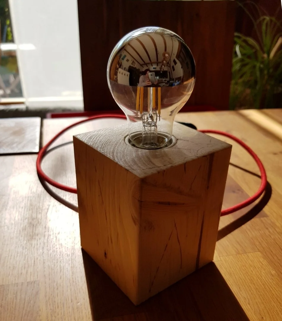 Eine Glühbirne auf einem Holzwürfel, mit einem roten Kabel im Hintergrund und einer Pflanzen im Raum.