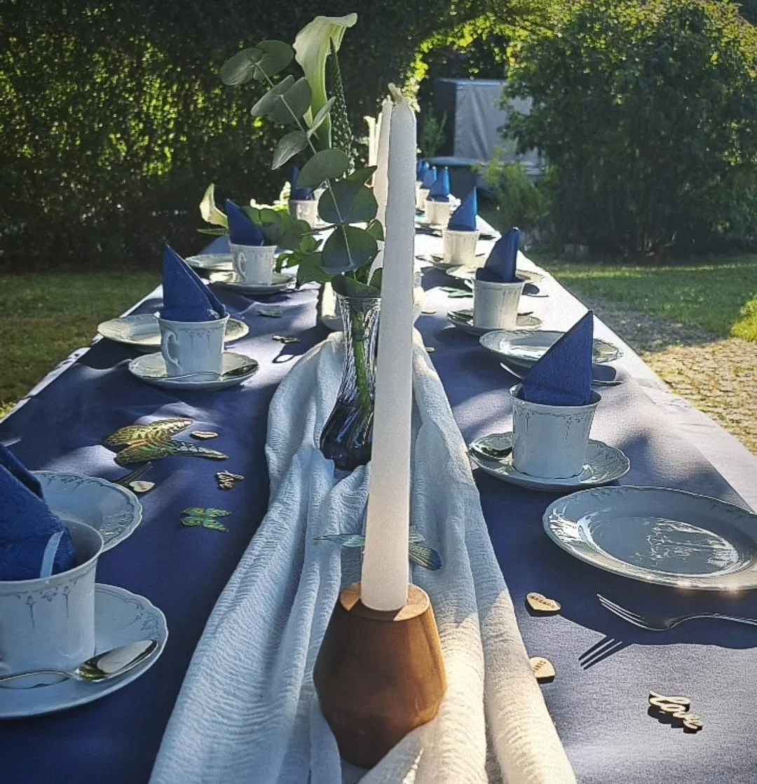 Ein langer Gartentisch ist festlich gedeckt mit weißen Tellern, weißen Kaffeetassen auf goldfarbenen Untertellern, blauen Stoffservietten, sowie Kerzen und Dekorationen in der Mitte, im Freien, umgeben von Bäumen, bei Sonnenlicht.