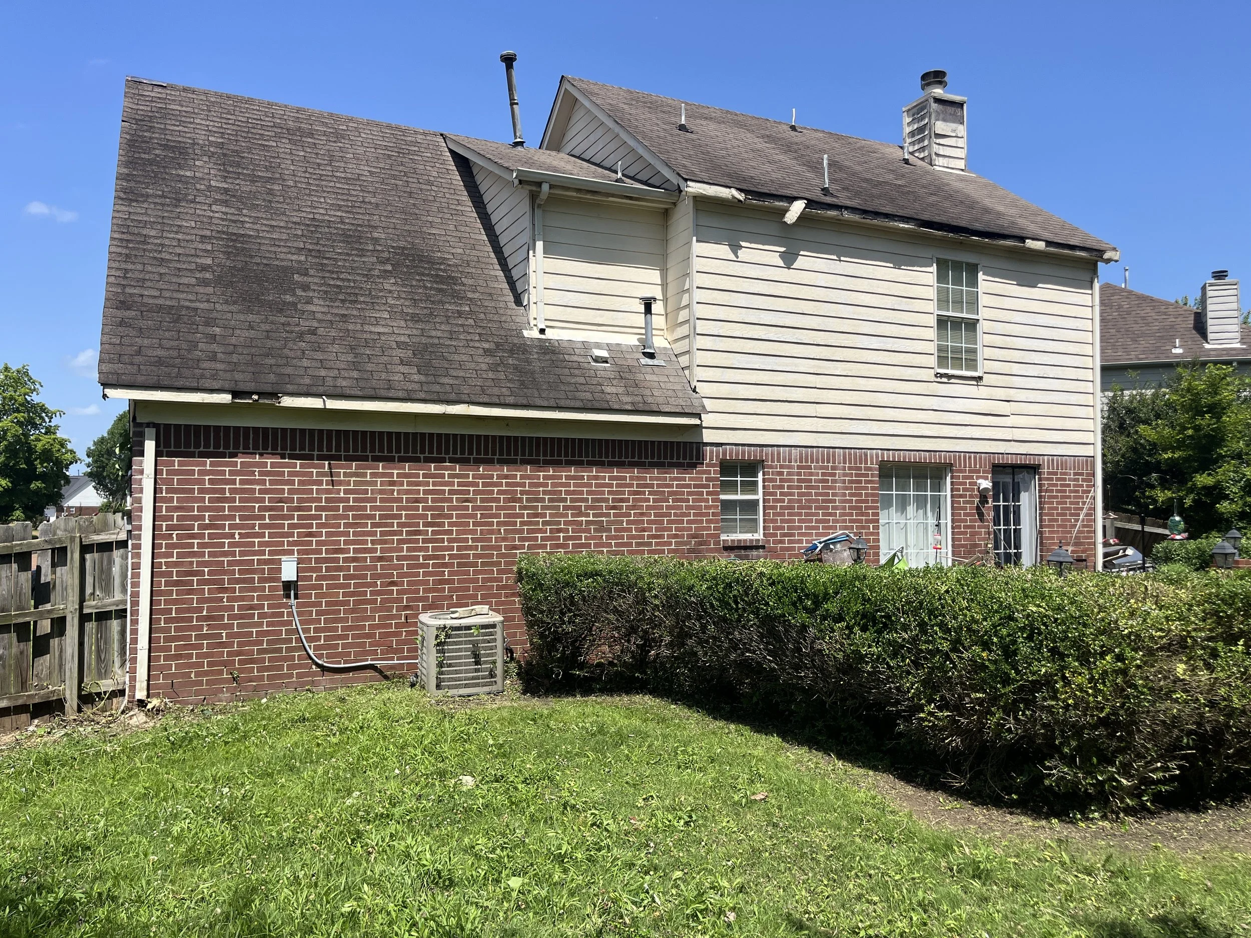 Rear view of a two-story house with a brick lower level and light-colored siding upper level. The house has multiple chimneys and a damaged section of the roof with a broken gutter. There is a small well-maintained garden and an air conditioning unit outside.