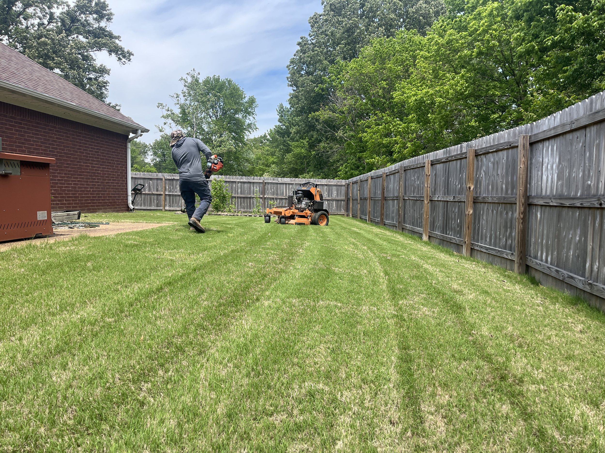 A person standing on a lawn, holding a leaf blower, working near a wooden fence with trees in the background.