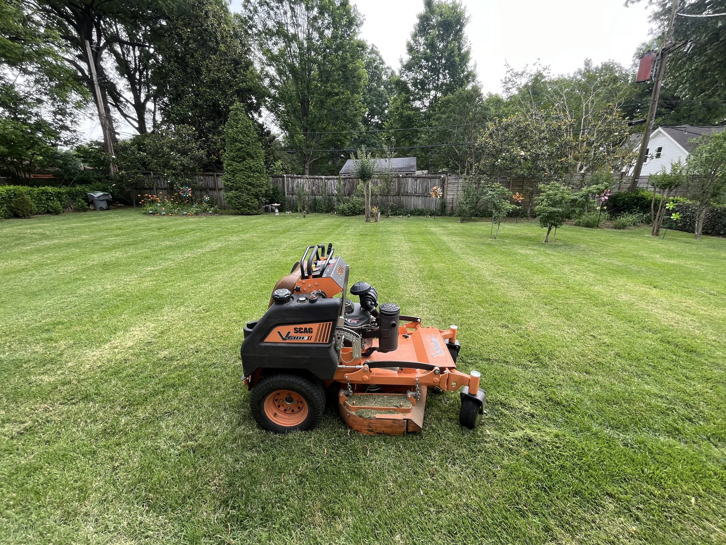 A backyard with a well-maintained green lawn, trees, flowers, and a wooden fence. There are decorative flower-shaped wind spinners and a white house visible in the background. In the foreground, there is an orange lawn mower with black control handles.