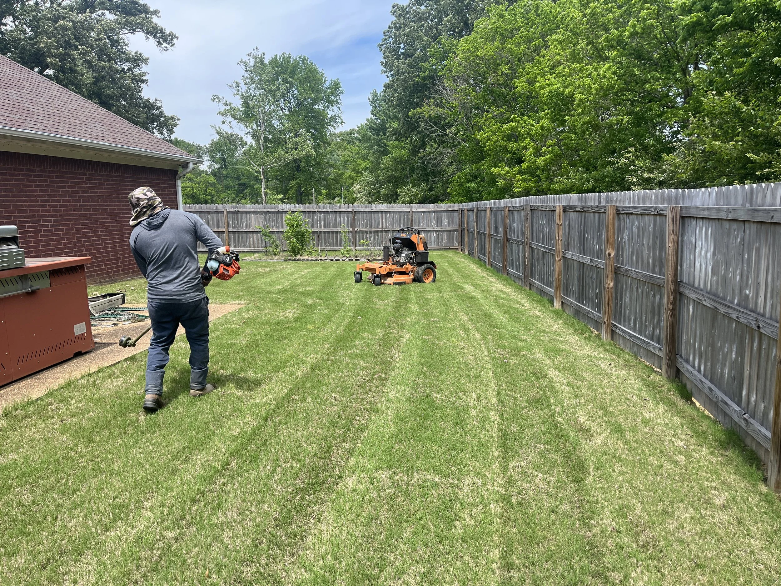 A person holding a leaf blower in a backyard, with a lawn mower nearby, a brick house to the left, and a wooden fence surrounding the yard. There are trees and a blue sky in the background.