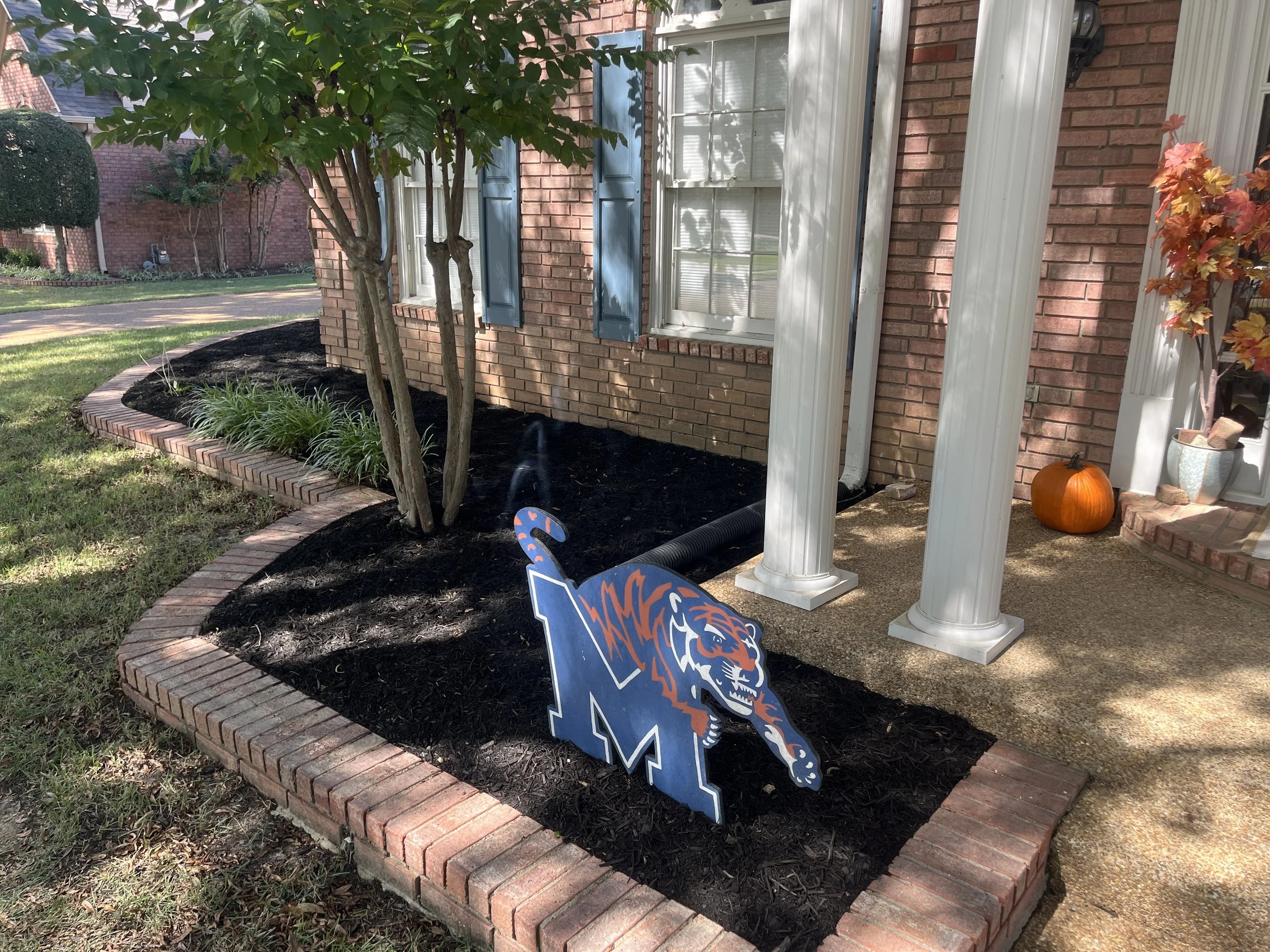 A brick house with blue window shutters and a porch, decorated with pumpkins and a National Football League sign, with trees and a lawn in the background.