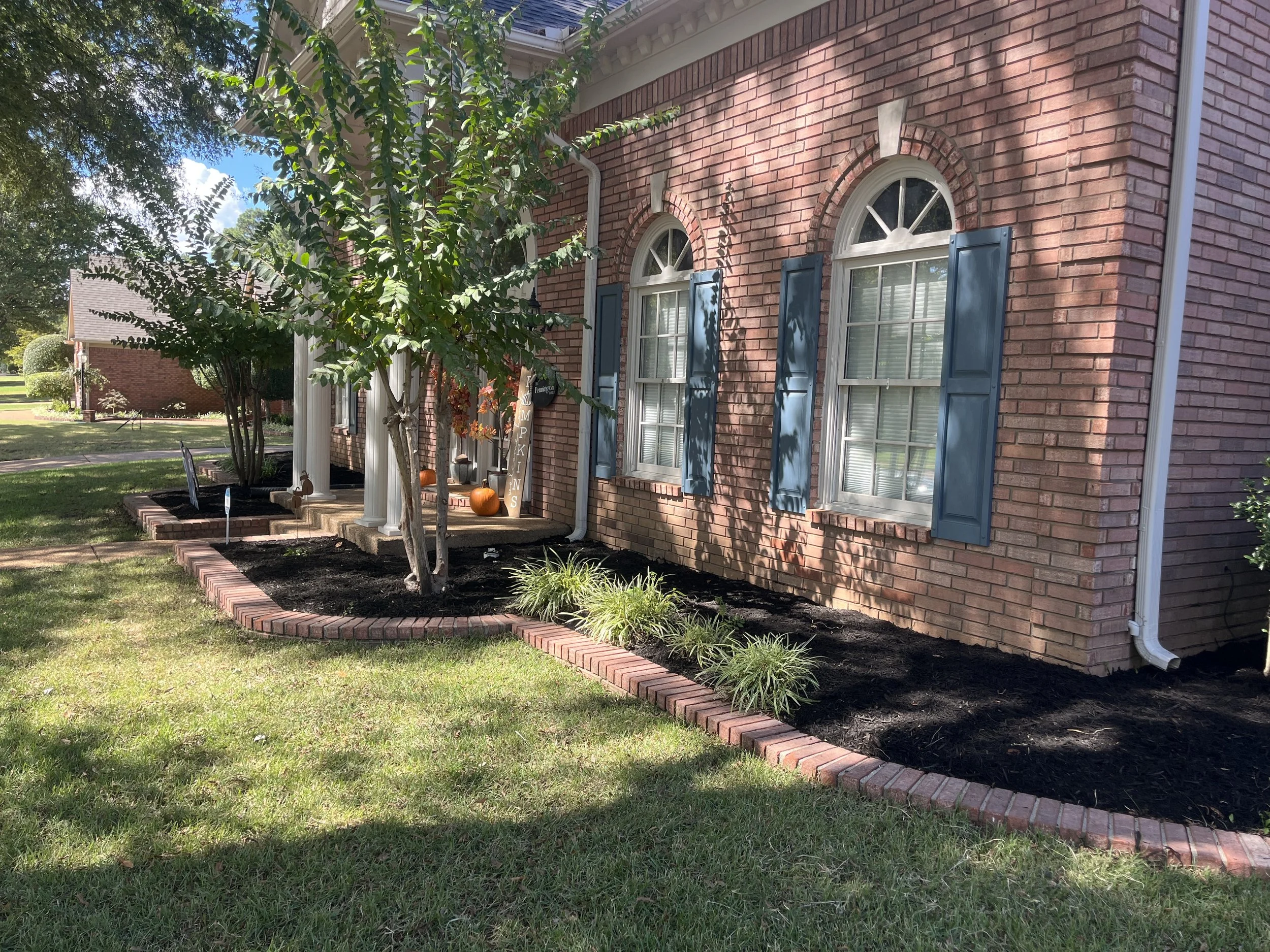 A brick house with three arched windows with blue shutters and a porch decorated with pumpkins and fall decorations. In front, there is a garden with a tree and a brick-edged flower bed.