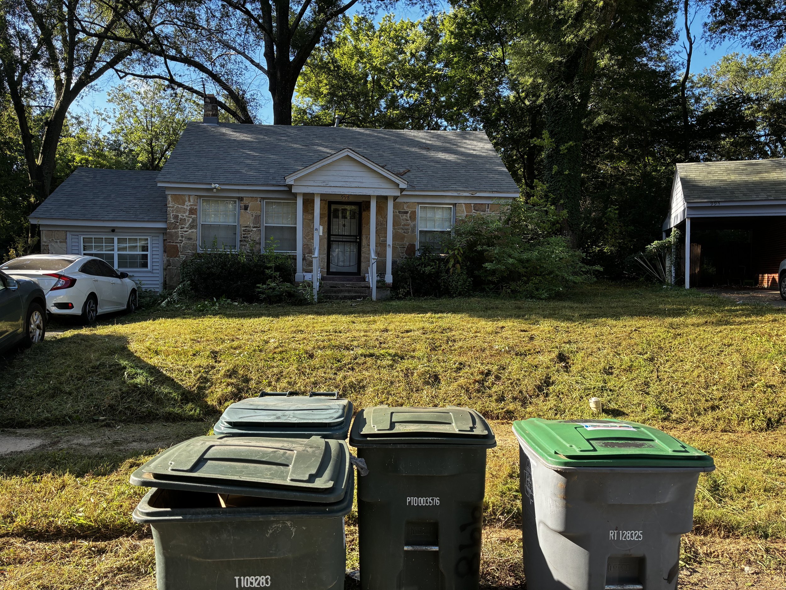 A house with a stone exterior and a front porch, beside two cars parked on the lawn, and three trash cans in the foreground, with trees in the background.