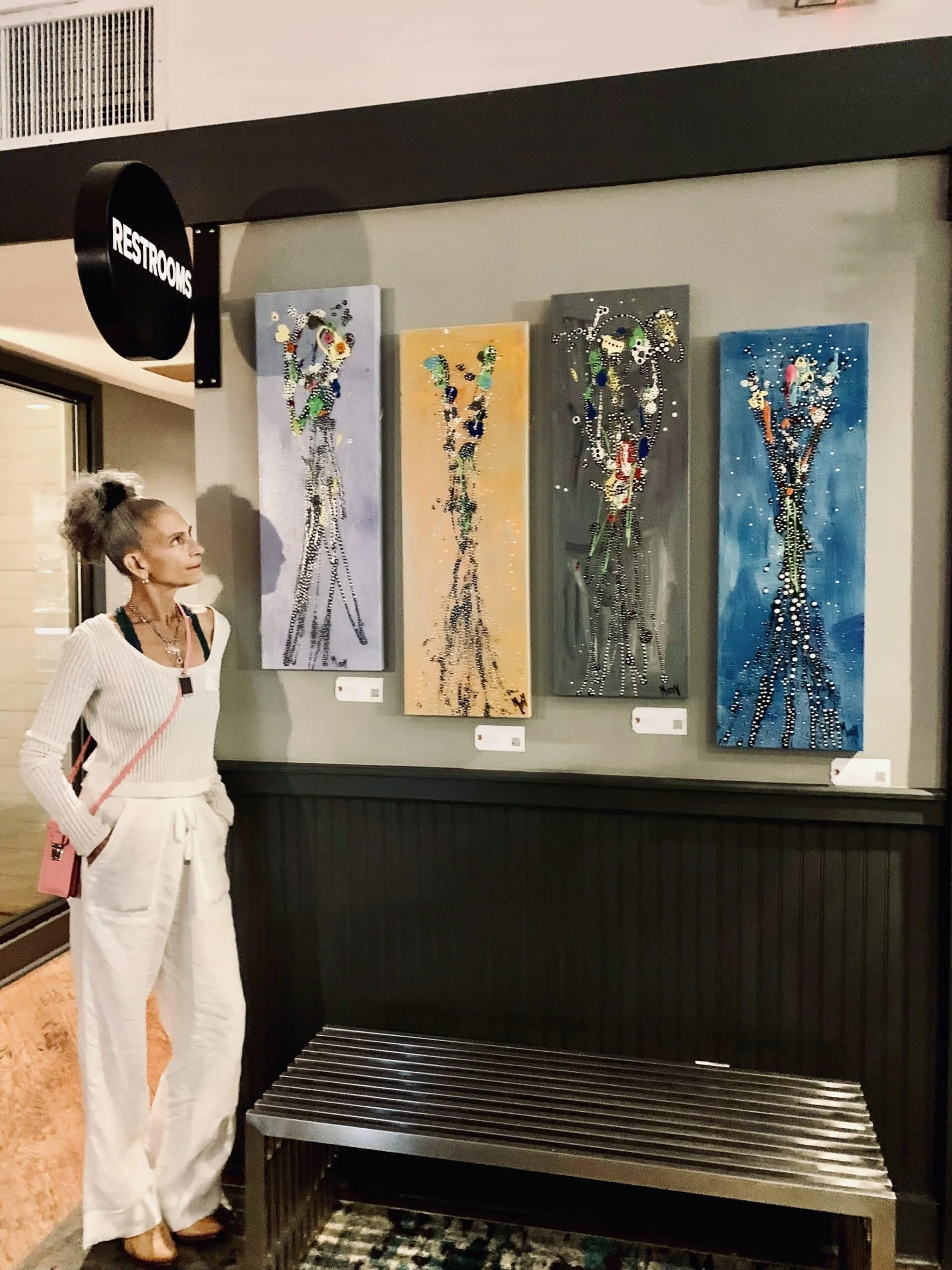 A woman with gray hair and light skin standing in front of four colorful abstract giraffe paintings in an art gallery or cafe, with a bench and a sign indicating restrooms nearby.