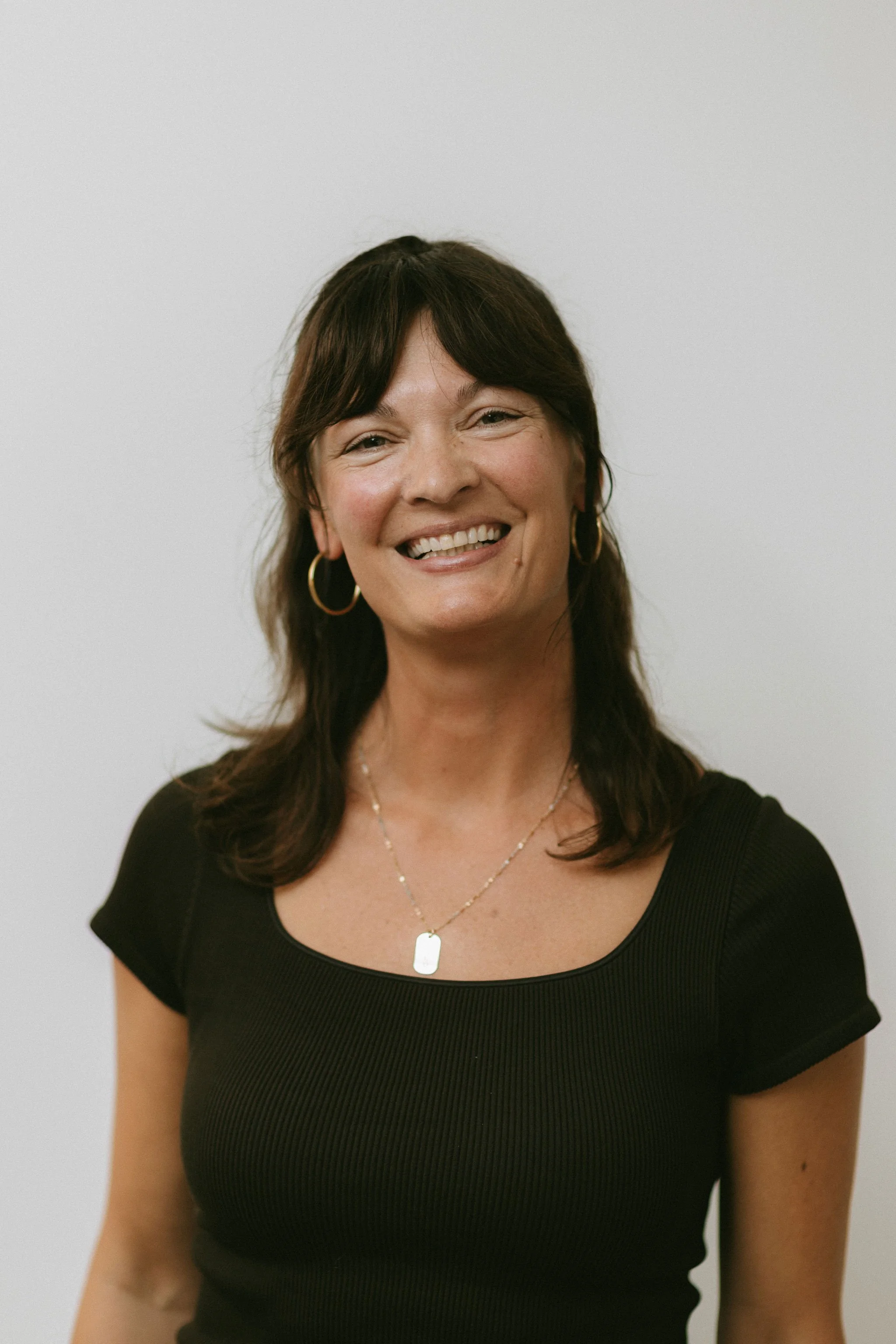 A woman with medium-length dark hair, wearing a black top, gold hoop earrings, and a gold necklace, smiling in front of a plain background.