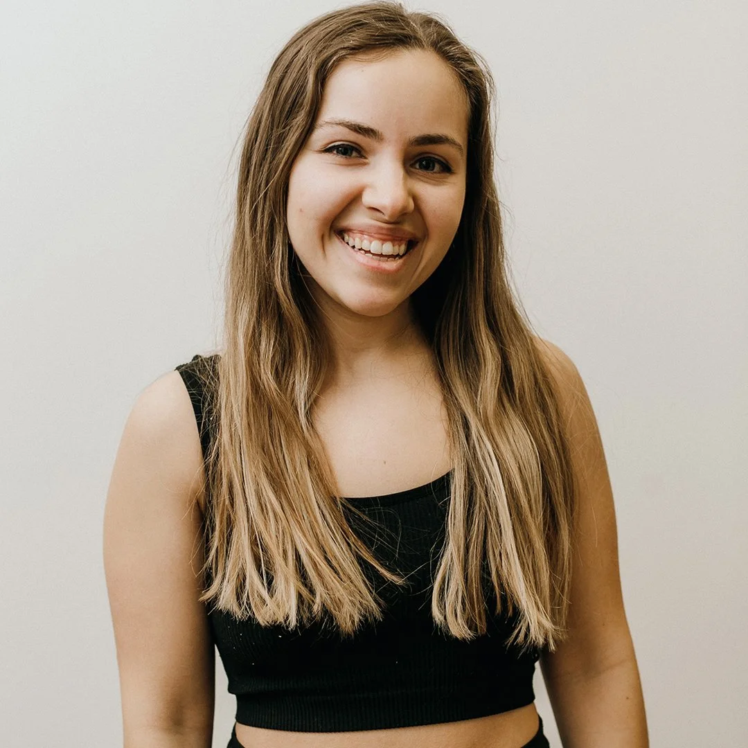 Smiling person with long hair wearing a black sleeveless top against a neutral background.