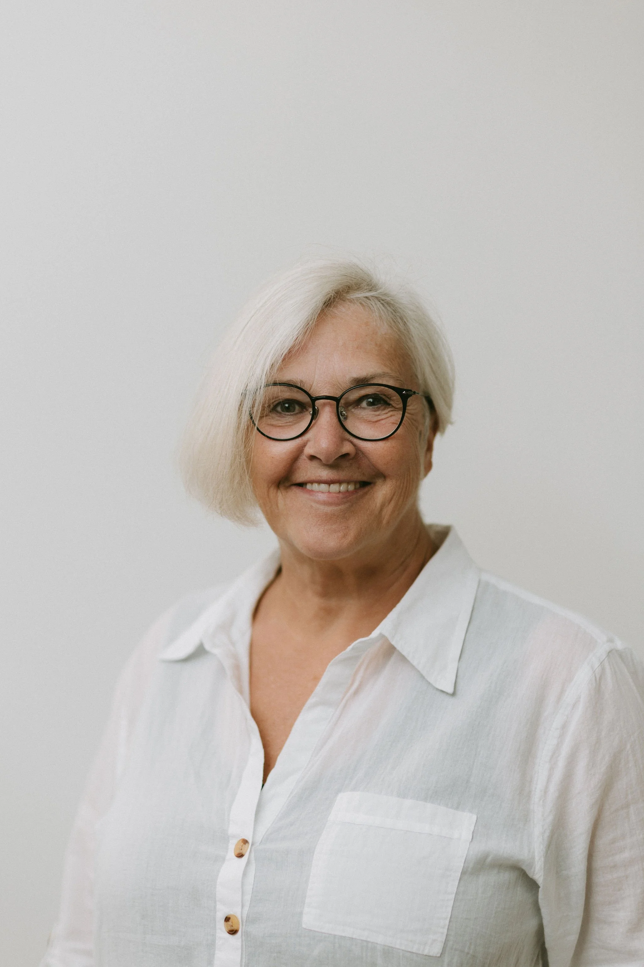 Older woman with short white hair and glasses smiling, wearing a white blouse, against a plain background.