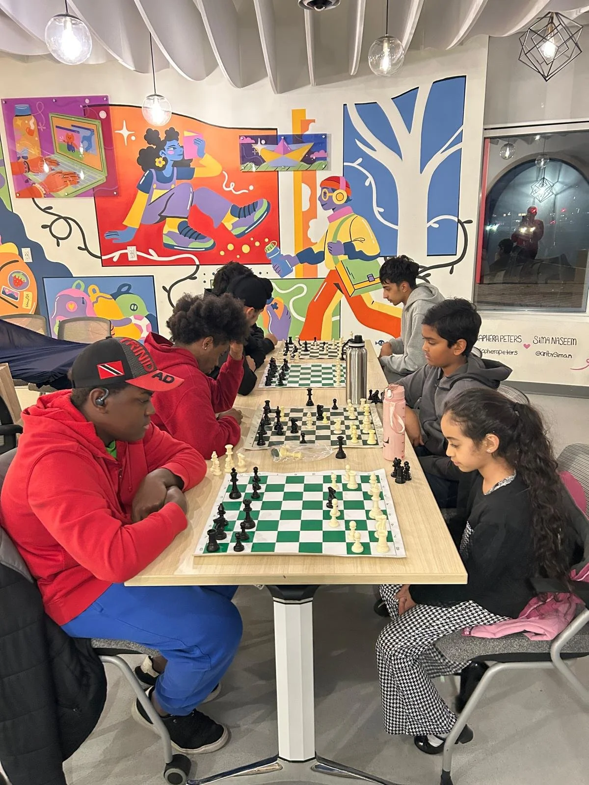 Children and teens play several chess games at a long table during an EnRoute 2 Success chess session, with chess boards and pieces arranged between them.