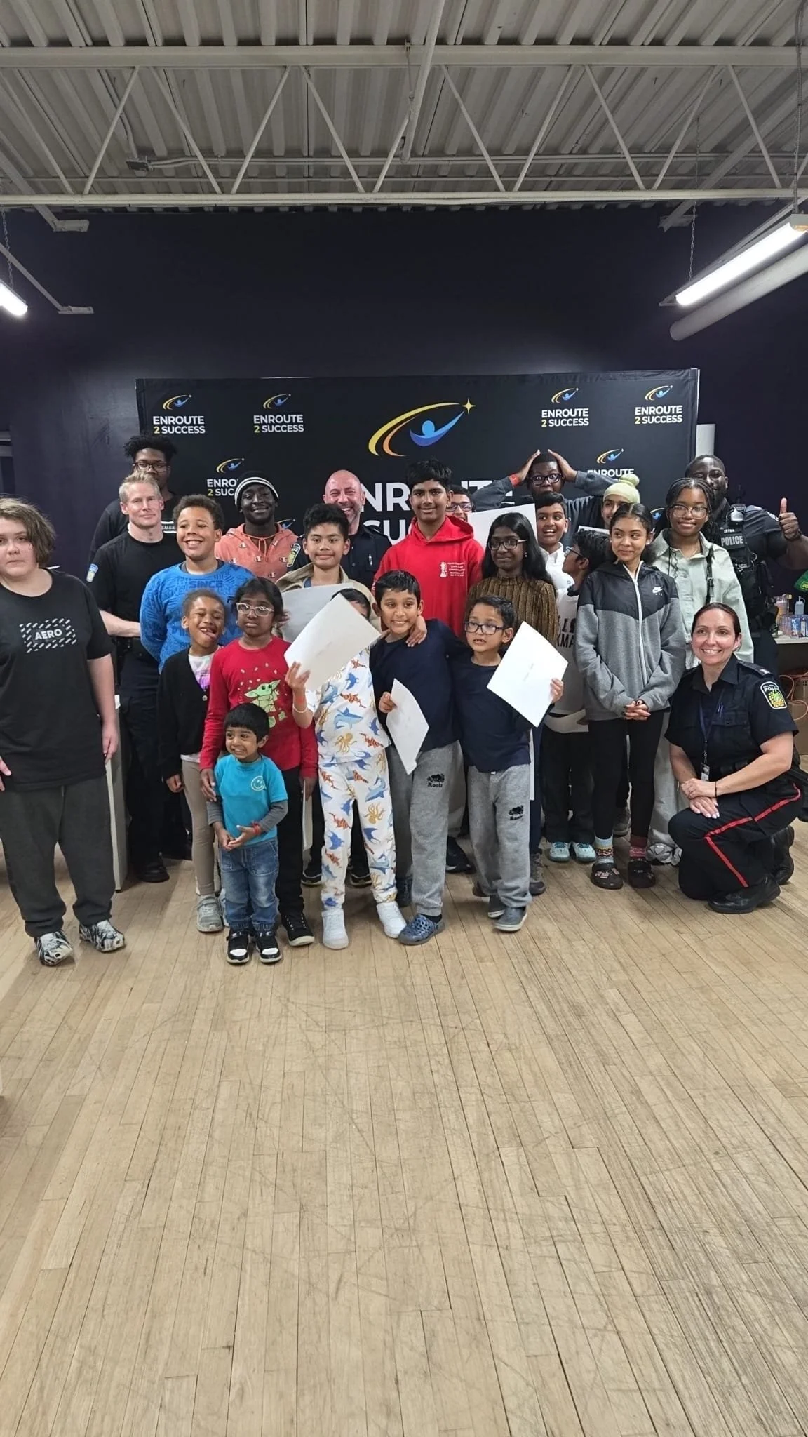 Group photo of youth, volunteers, and police officers posing together in front of an EnRoute 2 Success backdrop. Several youth hold certificates while others smile and celebrate indoors.