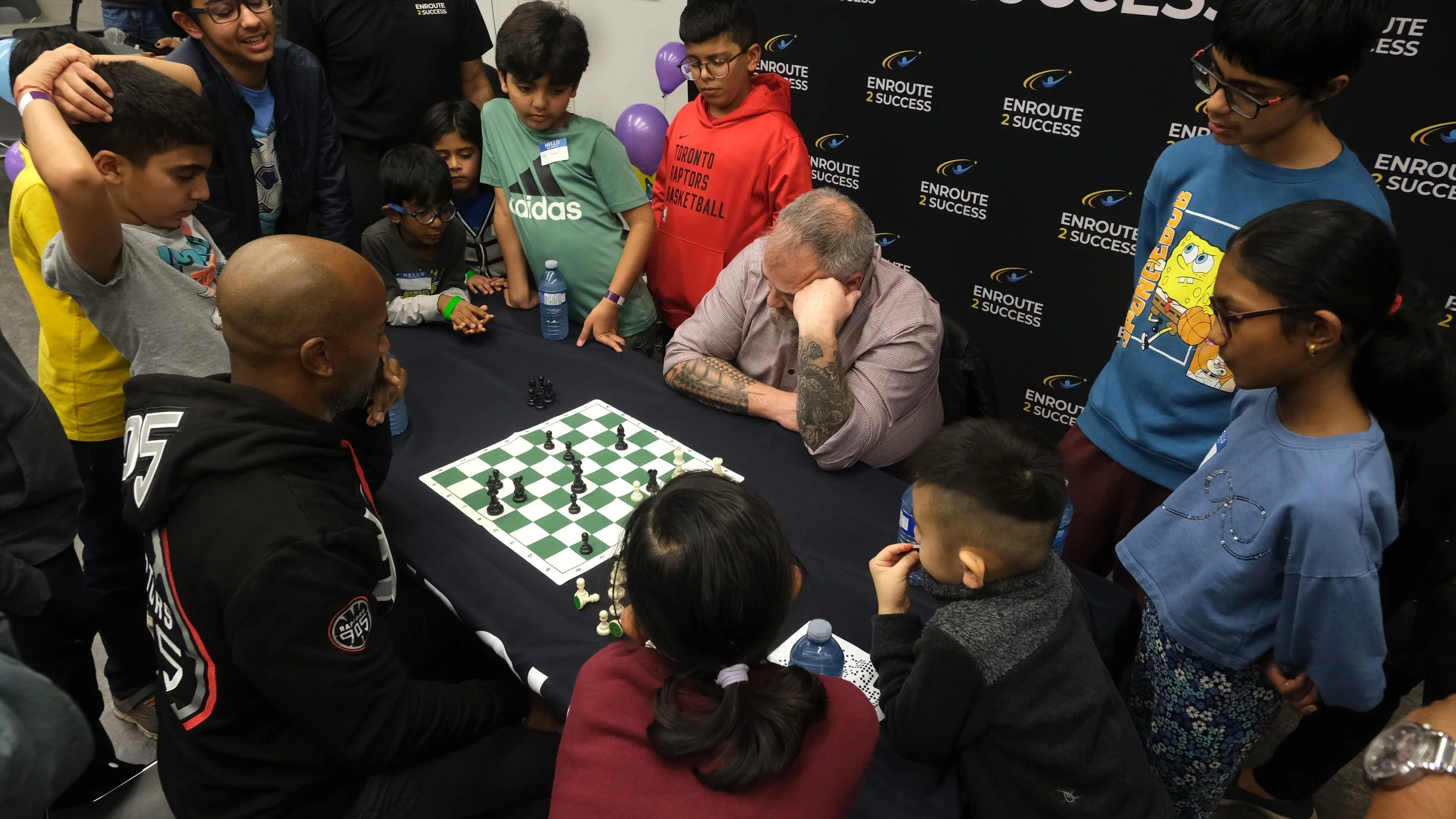 Youth gather around a chess board during a live match at an EnRoute 2 Success community chess event