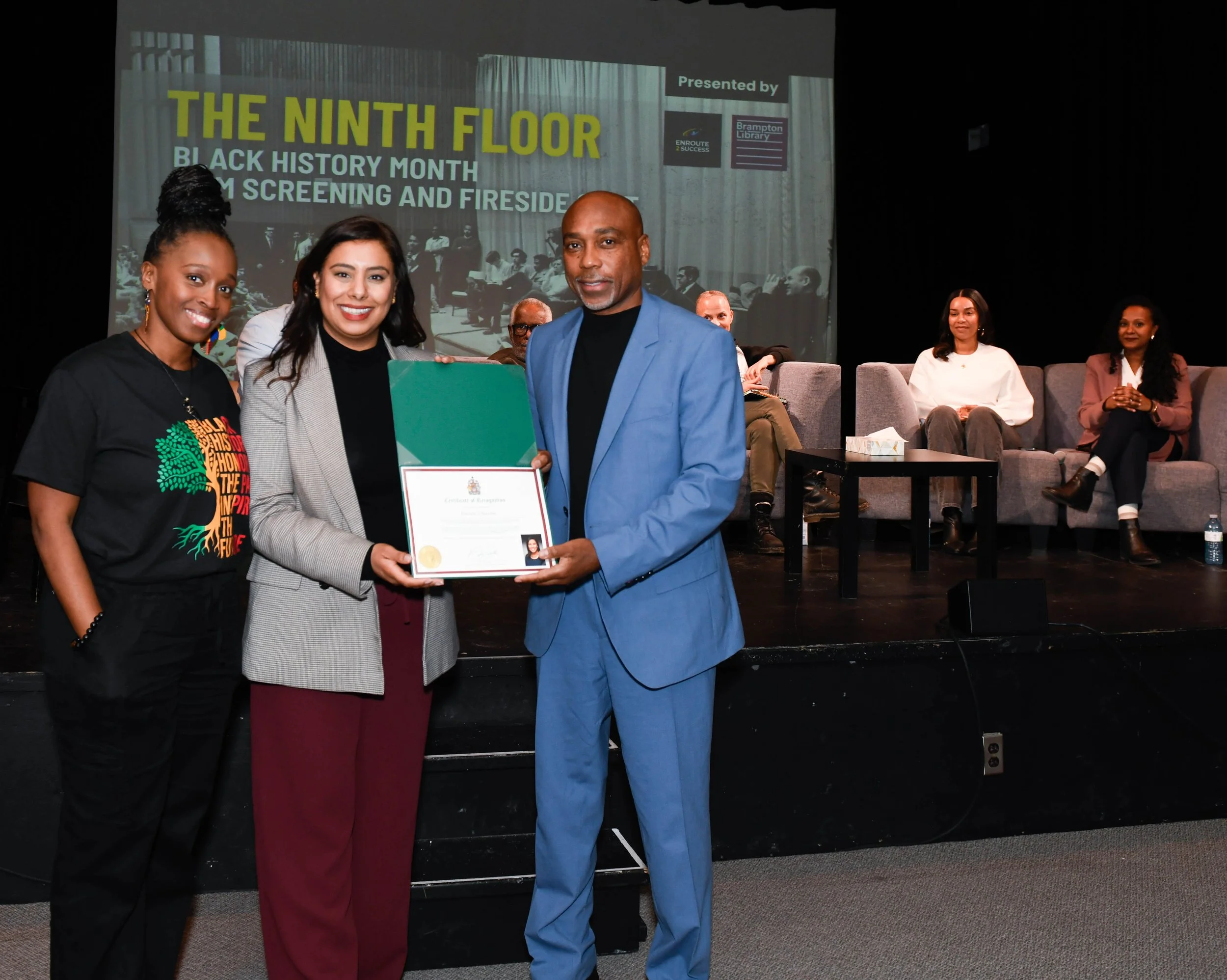 Three individuals stand at the front of a stage during an Enroute 2 Success event, holding a certificate and smiling toward the camera. A screen behind them shows “The Ninth Floor” alongside details of a film screening and fireside discussion, with p