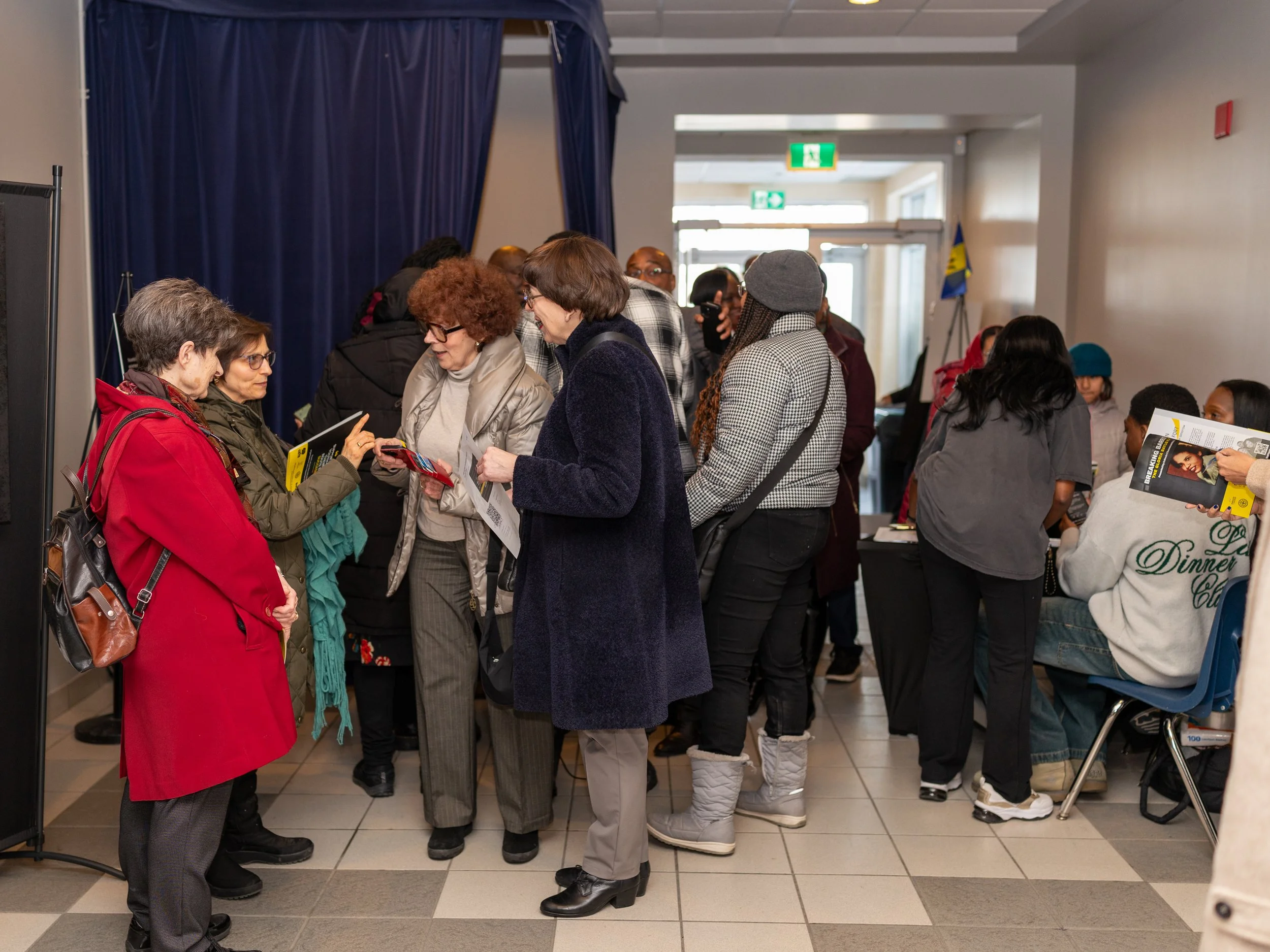 Attendees gather inside a venue during an Enroute 2 Success community event, speaking with one another and holding programs near an entrance and check-in area, creating a welcoming and busy atmosphere.