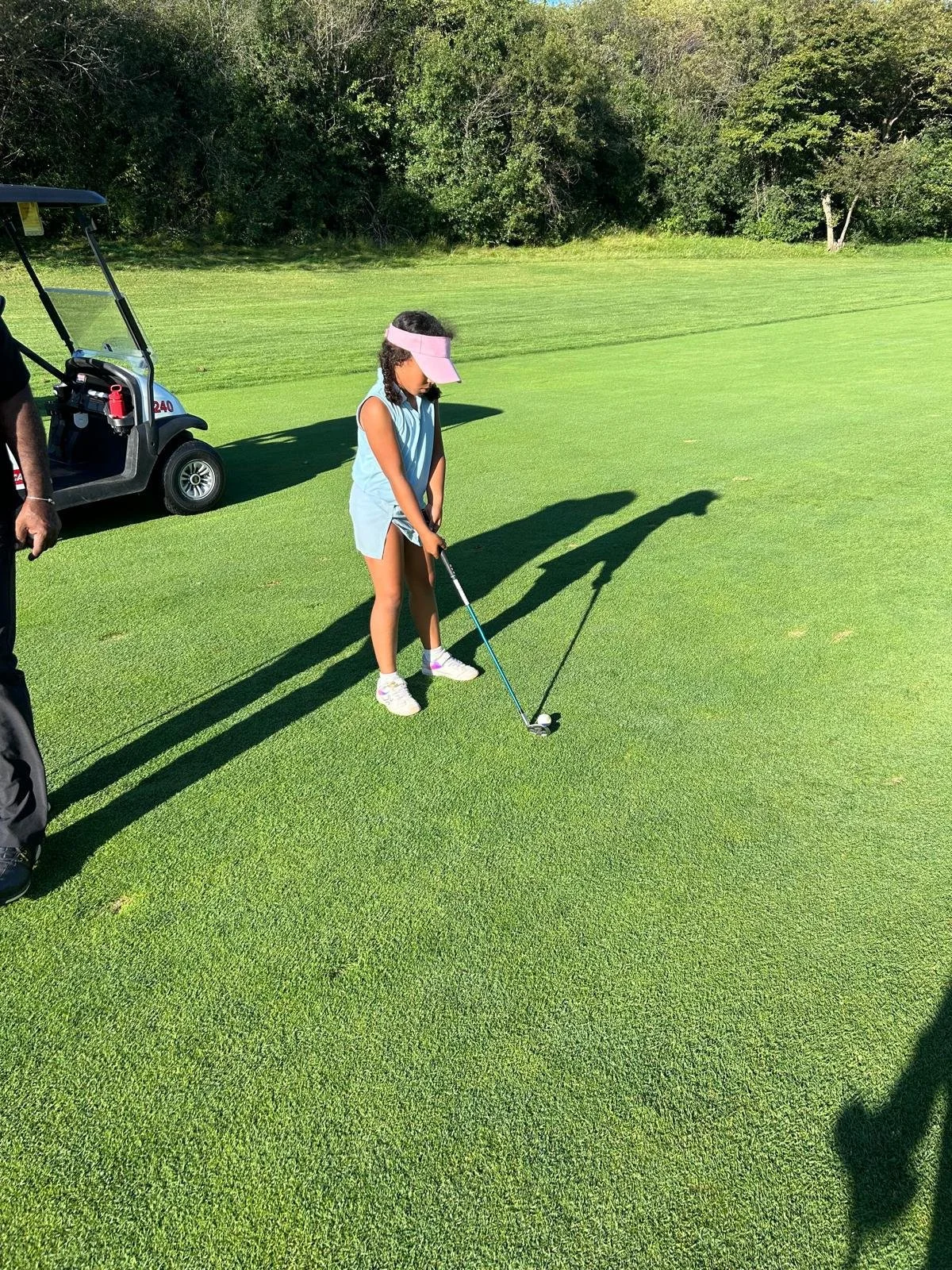 A young participant practices putting on a golf green during an EnRoute 2 Success golf session, concentrating on the ball while a coach stands nearby and a golf cart is visible in the background.