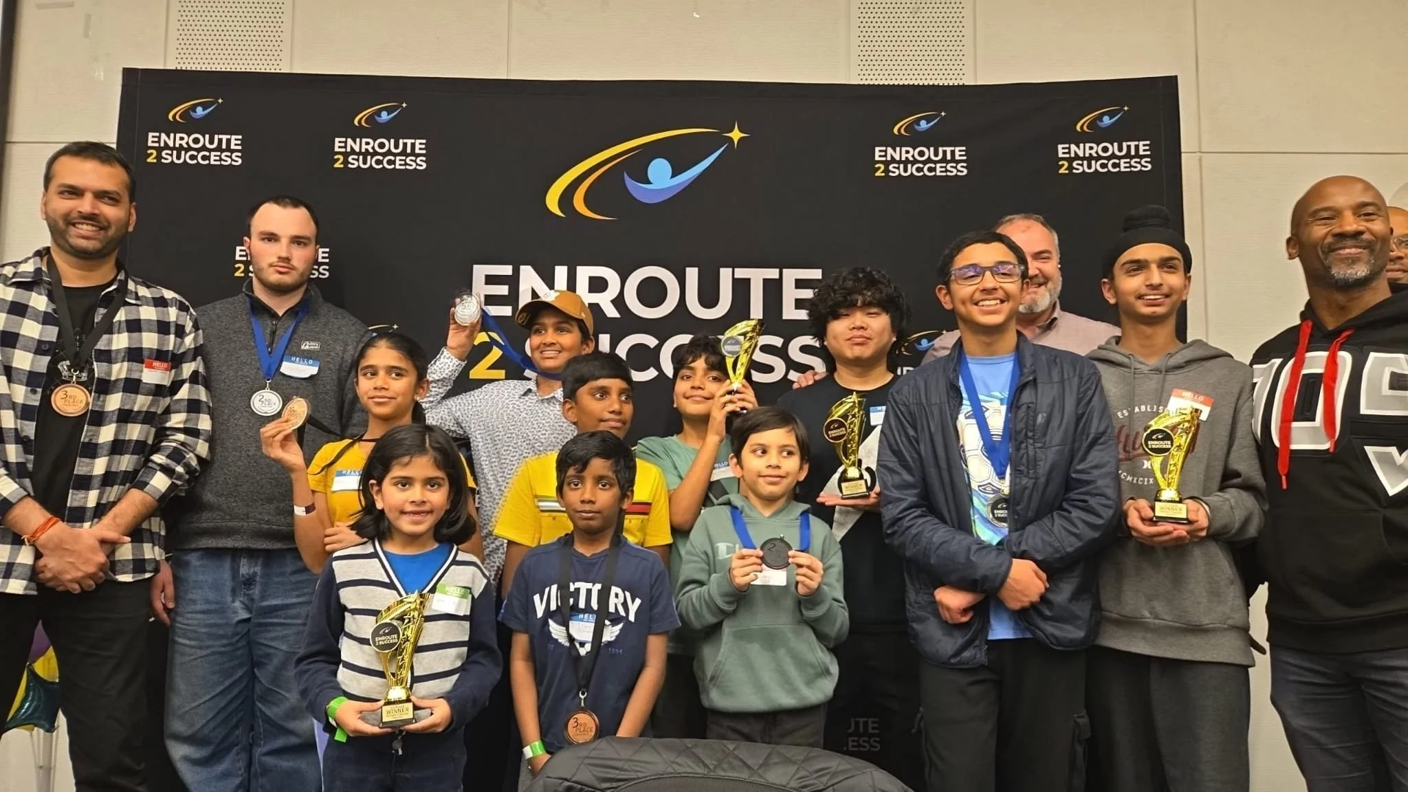 A group of children, teens and adults pose in front of an EnRoute 2 Success backdrop while holding chess trophies and medals after a chess event.