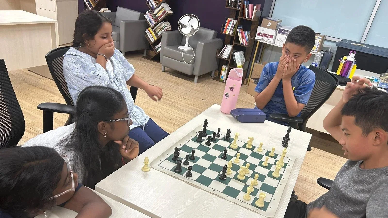 A group of youth participants gather around a chessboard during an EnRoute 2 Success session, reacting with surprise and excitement as they study a critical moment in the game in a classroom setting with bookshelves in the background.