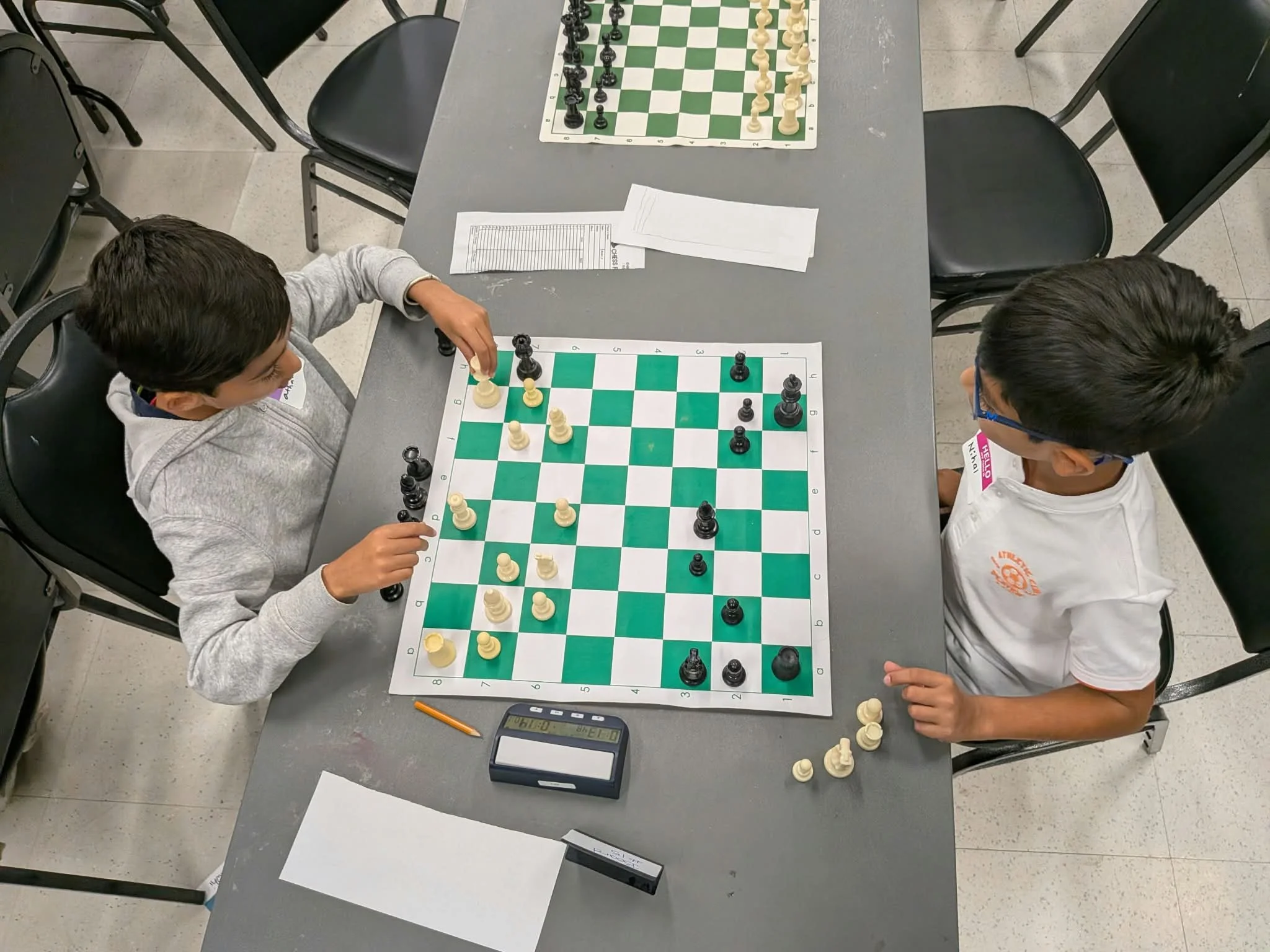 Overhead view of two youth seated across from each other during a chess match. One player reaches to move a piece while the other watches the board. A chess clock, captured pieces, and a pencil are visible on the table.