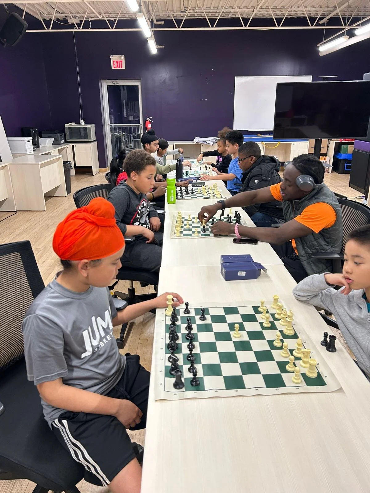 Children and teens play several chess games at a long table during an EnRoute 2 Success chess session, with chess boards and pieces arranged between them.