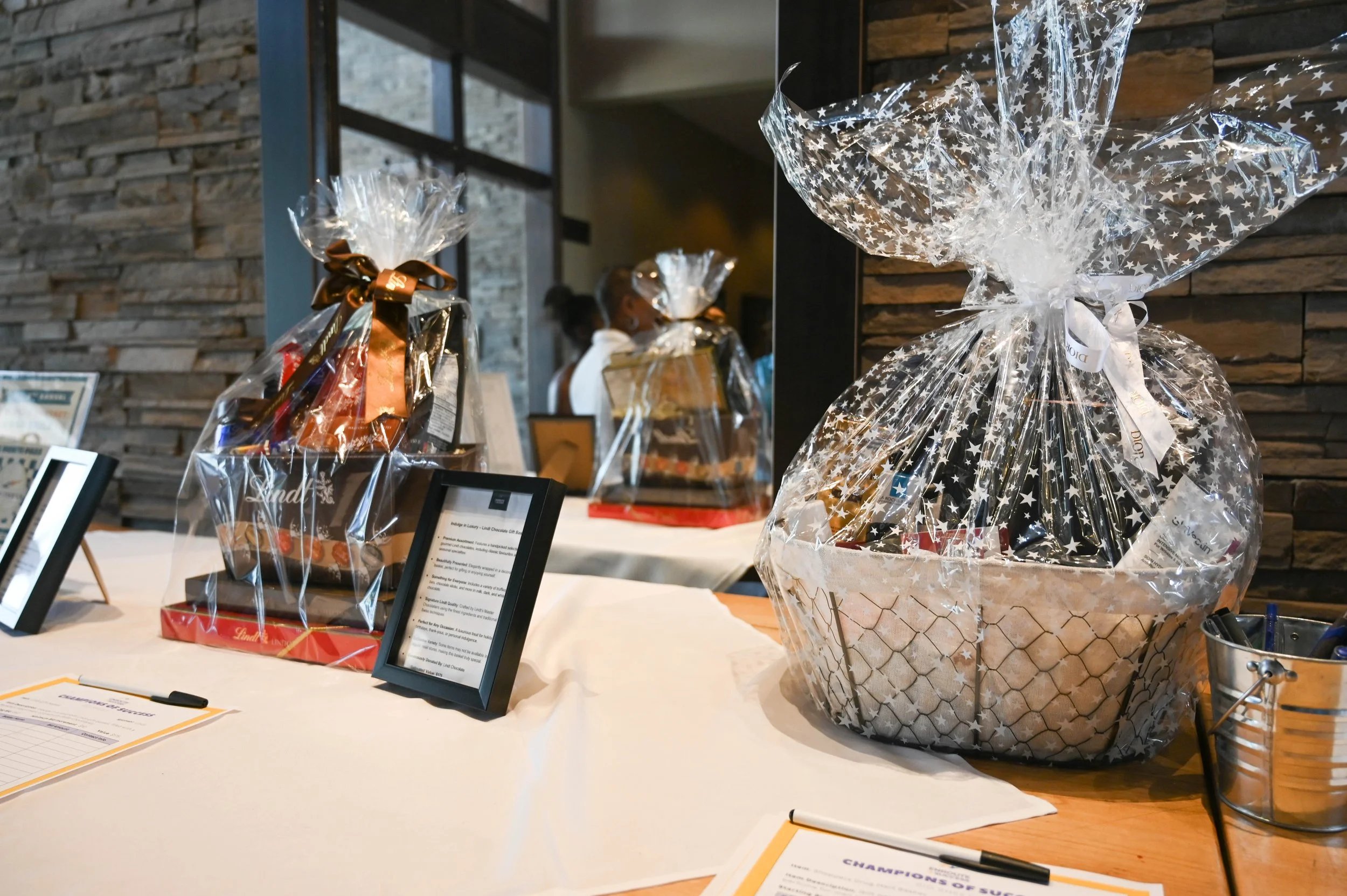 Silent auction table displaying wrapped gift baskets and prize packages for the Champions of Success Golf Tournament fundraiser, featuring raffle items and donation baskets arranged for guests and bidding participation.