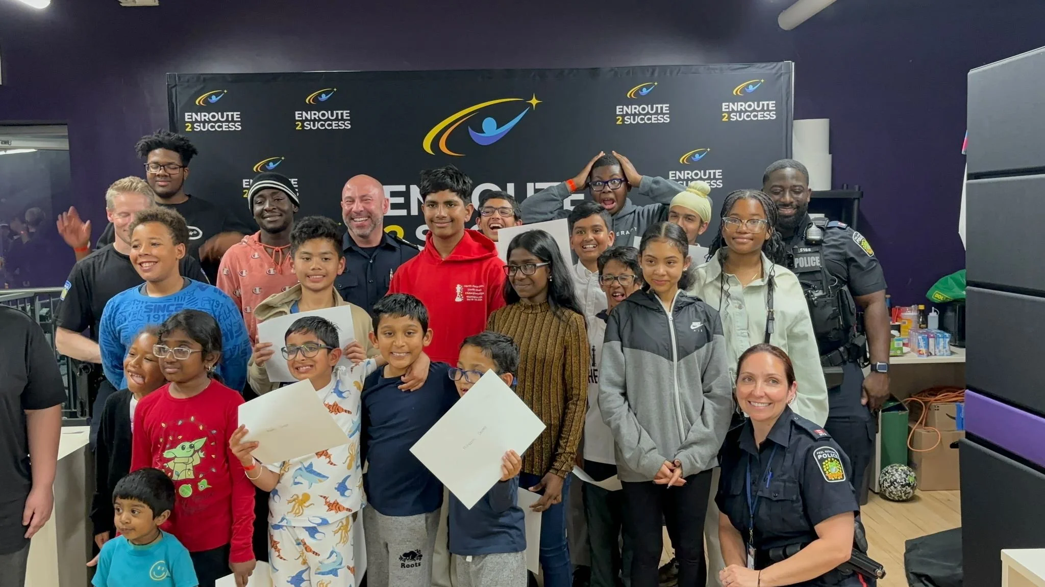 Group of youth participants and community members at an Enroute 2 Success event standing in front of the Enroute 2 Success backdrop, some holding certificates