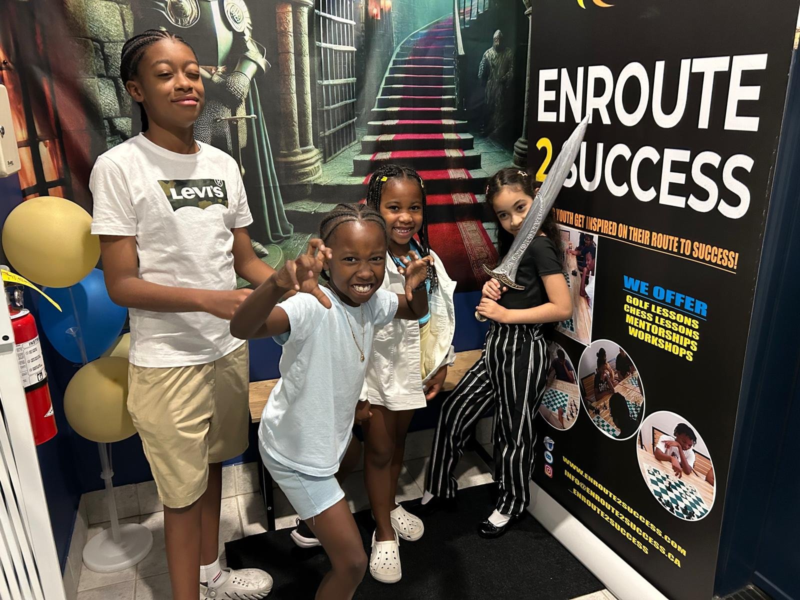 Four youth participants pose and smile in front of an EnRoute 2 Success banner at a community event, with one holding a toy sword and others playfully reacting, surrounded by balloons and promotional displays.